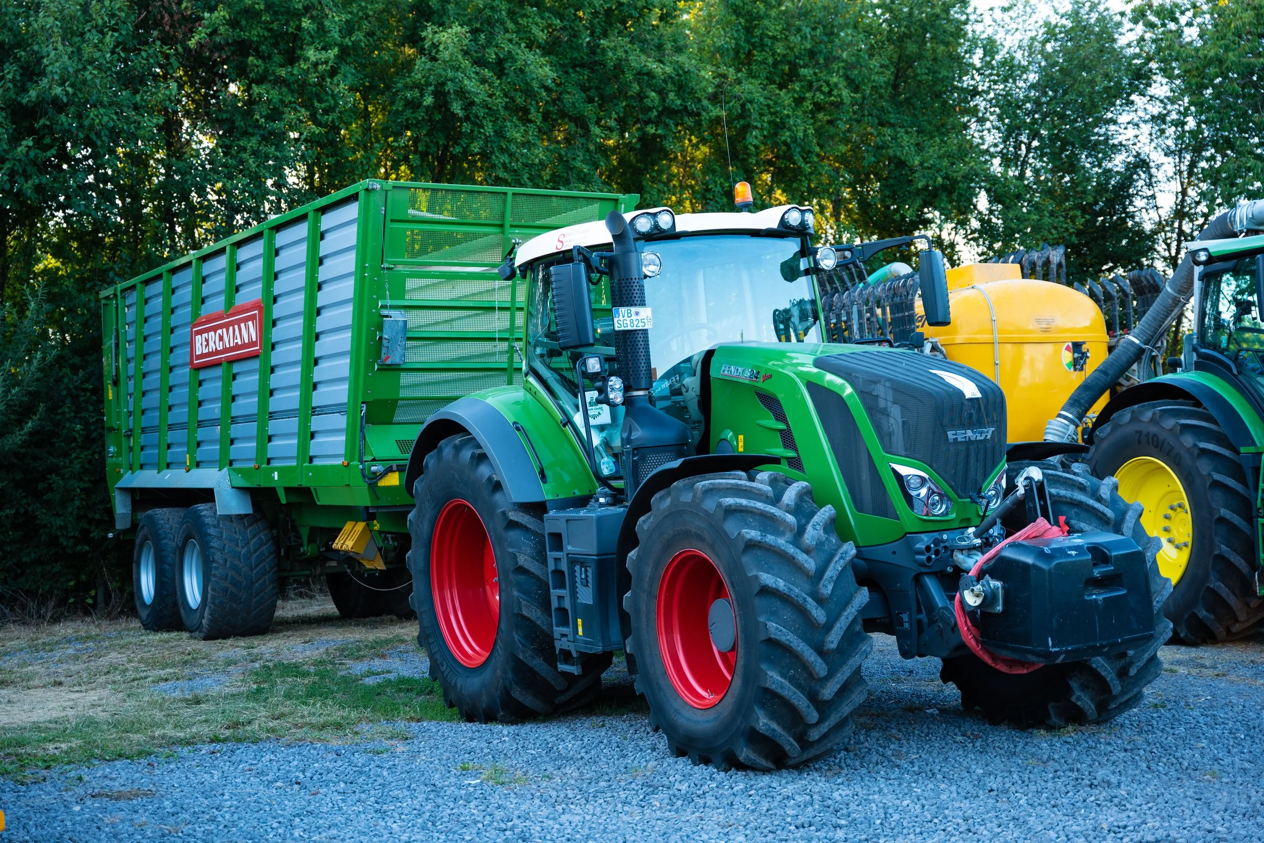 Ein grüner Traktor mit Anhänger, der das Bergmann-Logo trägt, steht auf einem Gelände mit Kies und Gras, umgeben von Bäumen.