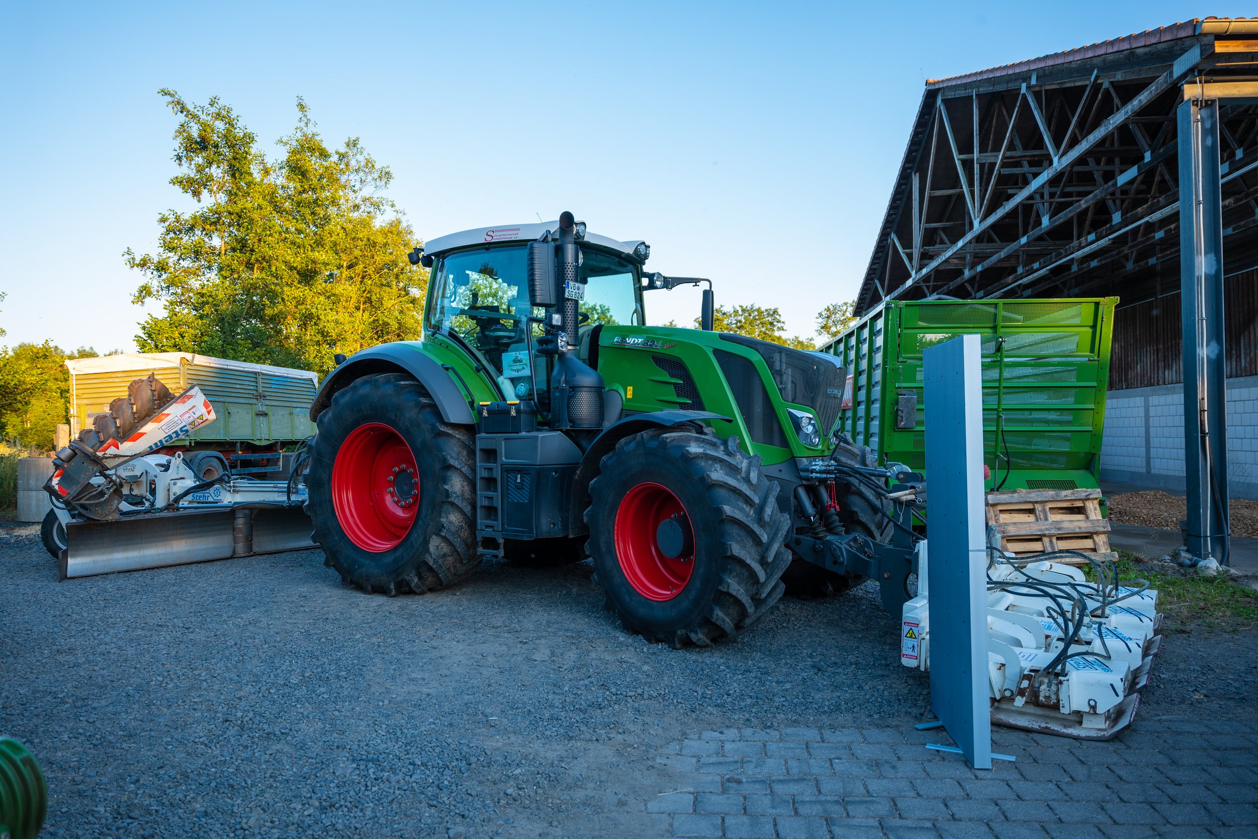 Grüner Traktor mit roten Rädern neben landwirtschaftlichen Maschinen und Anhängern auf einem Bauernhof.