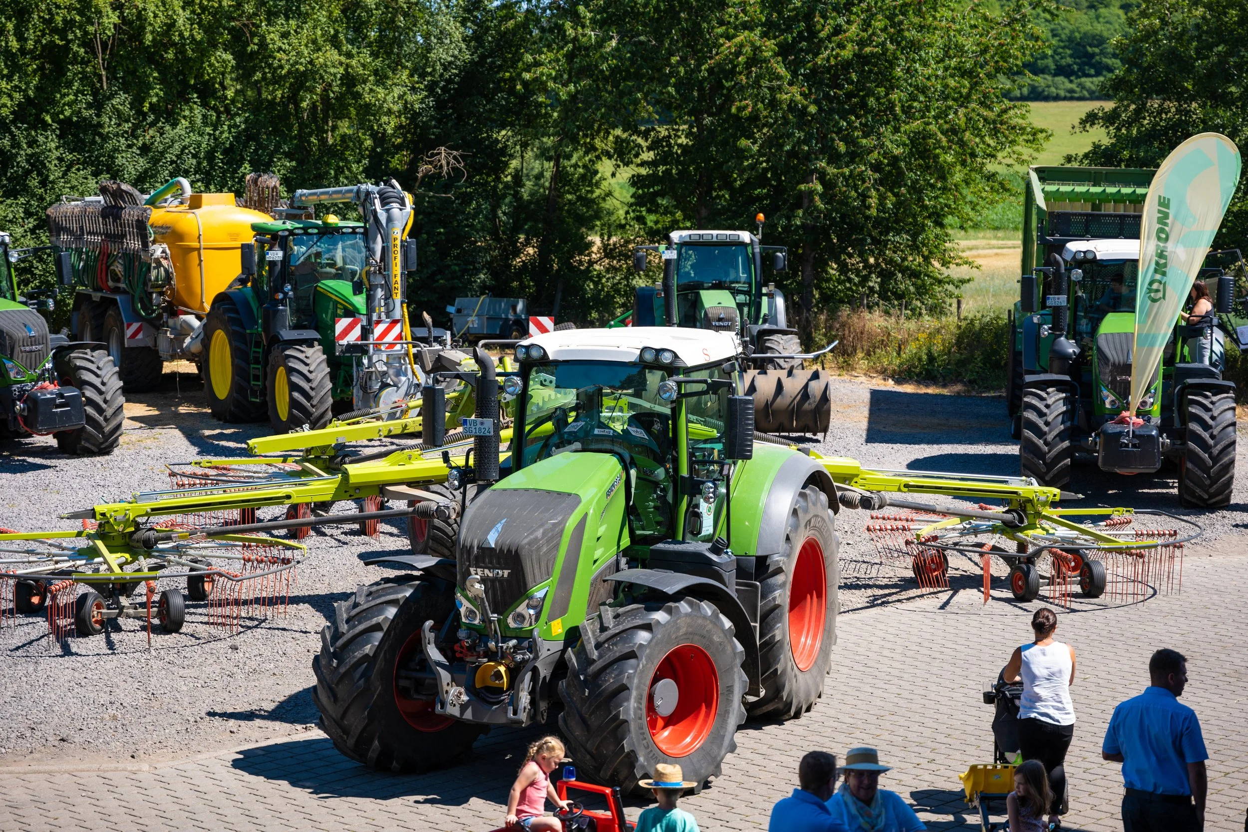 Landwirtschaftsausstellung mit mehreren Traktoren und landwirtschaftlichen Maschinen auf einem gepflasterten Gelände, besichtigt von Menschen. Im Hintergrund sind Bäume und ein Banner zu sehen.
