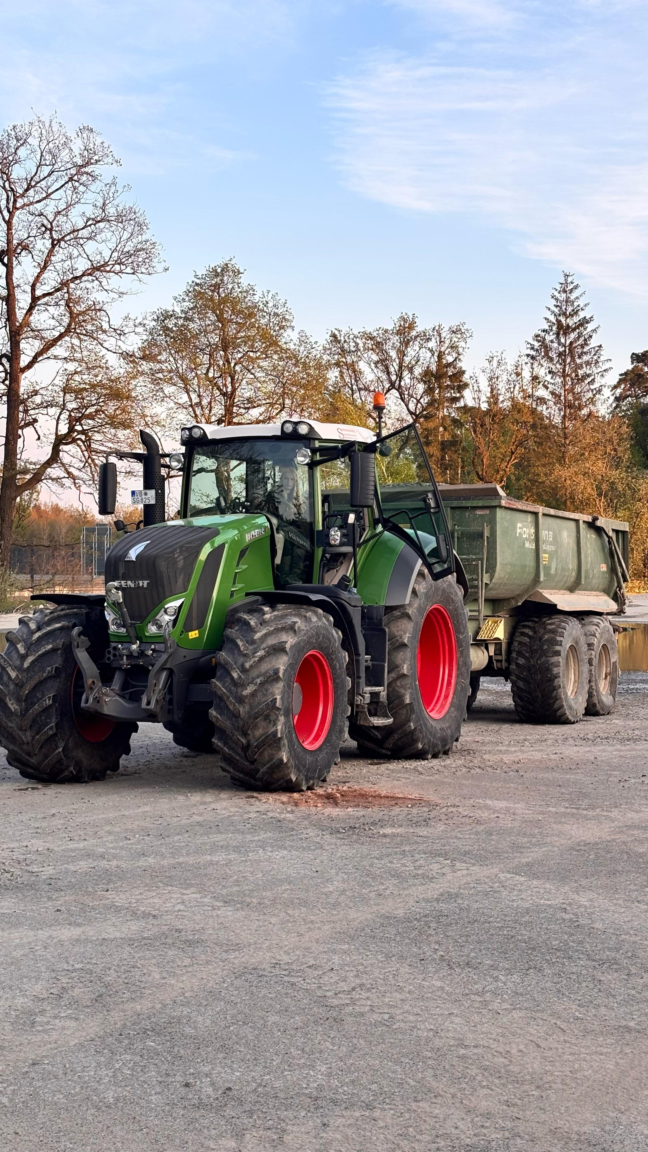 Traktor der Marke Fendt mit einem Anhänger auf einem Kiesplatz, umgeben von Bäumen und blauem Himmel.