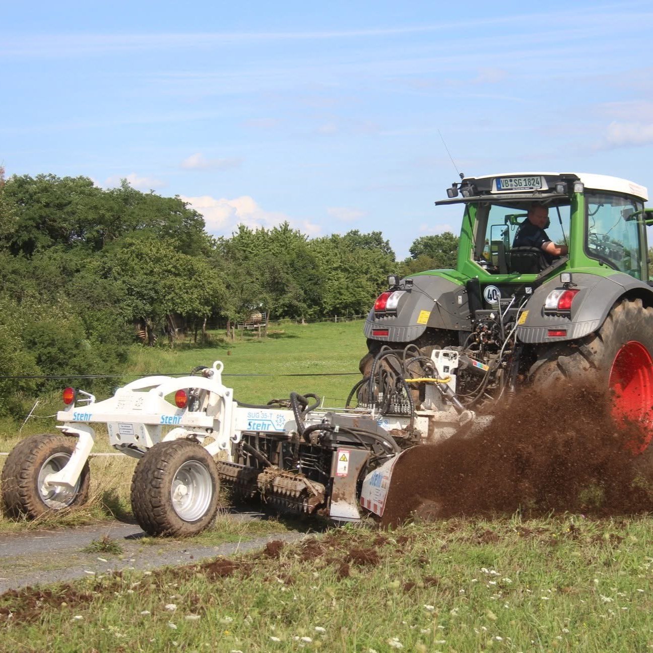 Traktor mit angebauter Bodenfräse, der eine Erdoberfläche auf einem Feld bearbeitet. Im Hintergrund sind Bäume zu sehen.