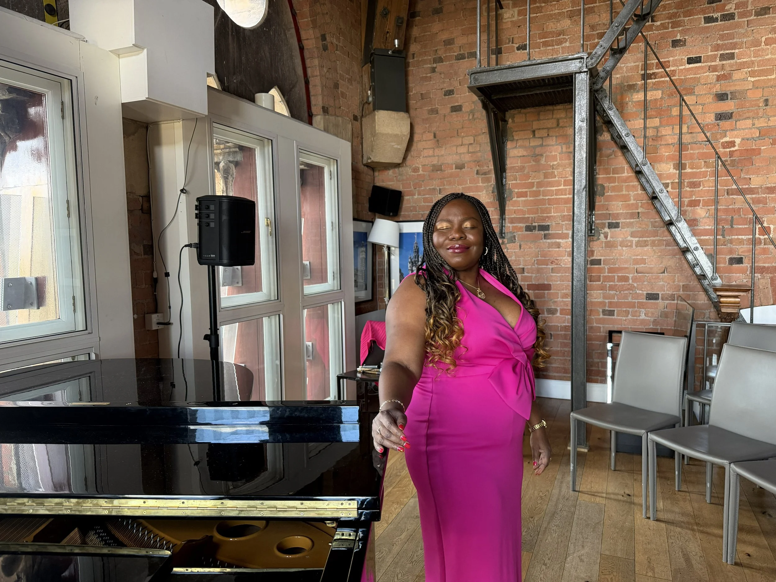 Victoria standing at the piano in the Clock Tower St Pancras. She is wearing a bright pink dress and gold eye shadow. Her braids fall over her shoulders and she is smiling softly.