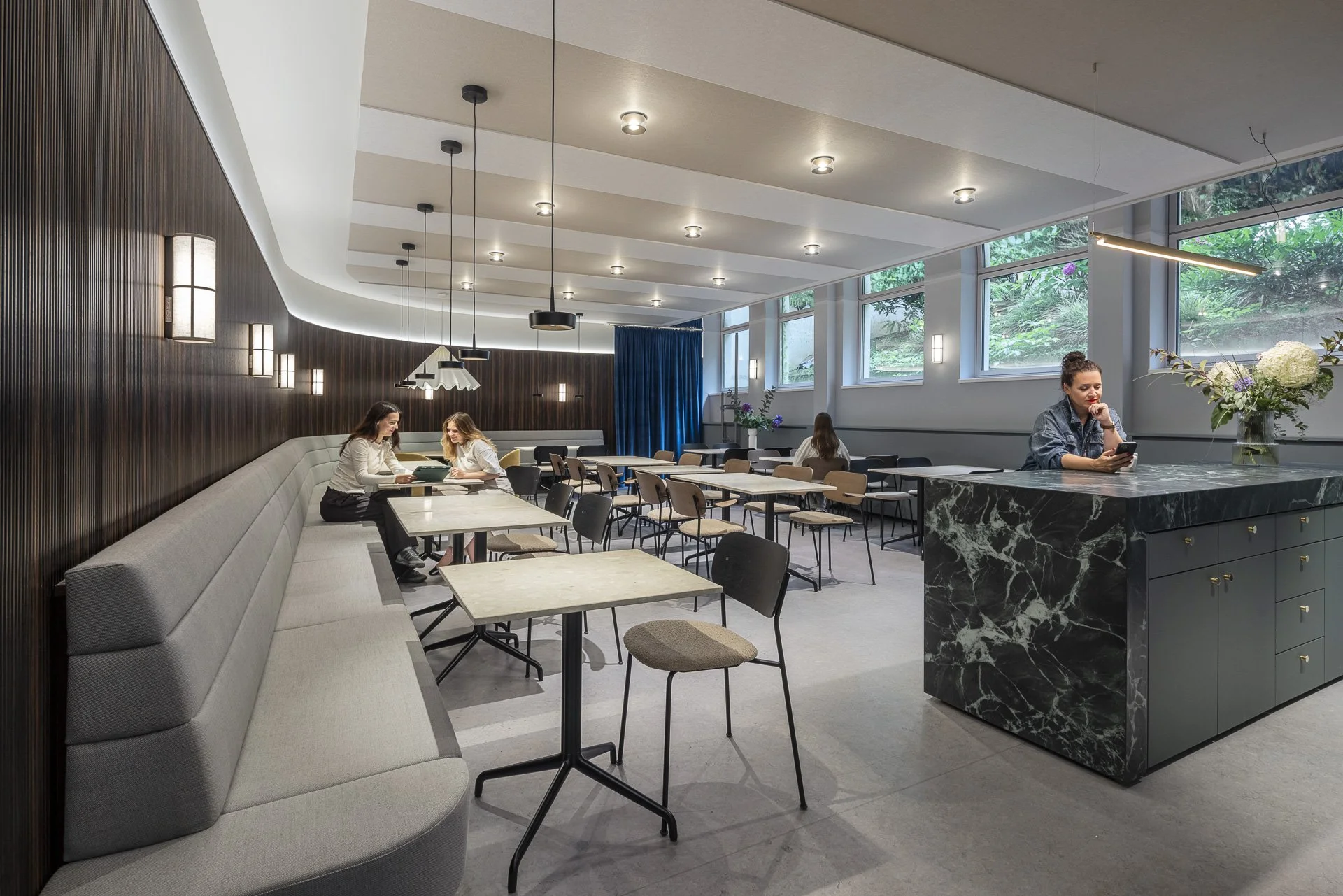 Café Gesis mehrere Stühle und Tische, an einem sitzen zwei Frauen, an der Bar sitzt Frau mit Smartphone