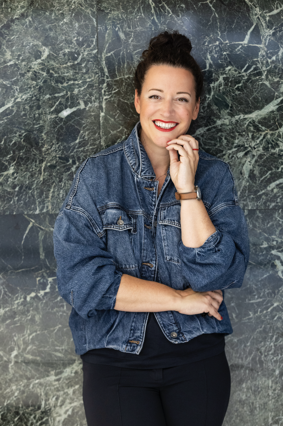 Frau in Jeansjacke, schwarzer Hose, hochgesteckte Haare lächelt, steht vor Wand aus Marmour