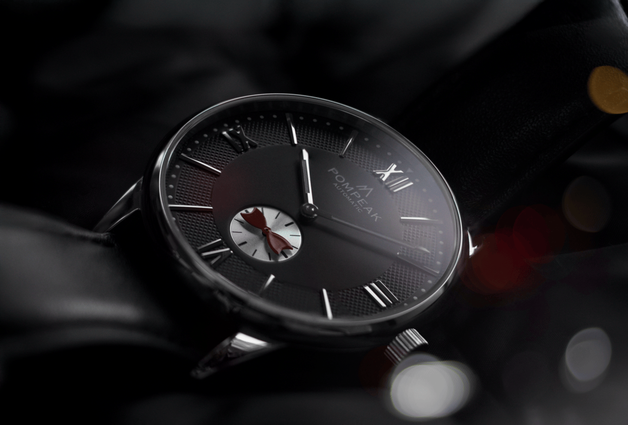 Pompeak Mallory Tux Edition watch featuring a black dial with textured outer ring, white sub seconds display, and distinctive bow tie seconds hand, shown on a leather strap in low light.