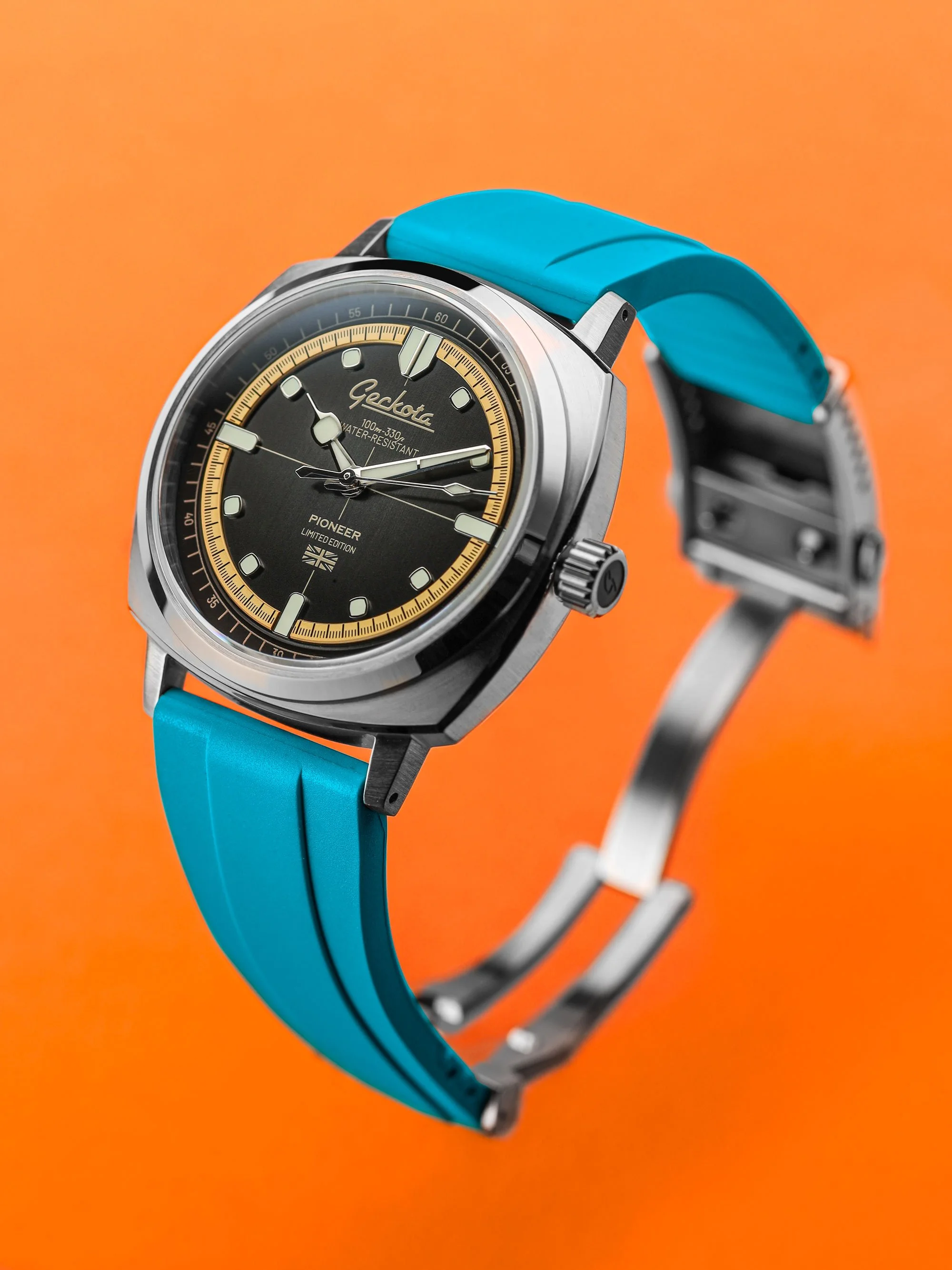 Geckota Pioneer Graphite Special Edition watch with a brushed grey dual layer dial, cushion shaped stainless steel case, and bright blue rubber strap, shown against a vivid orange background.