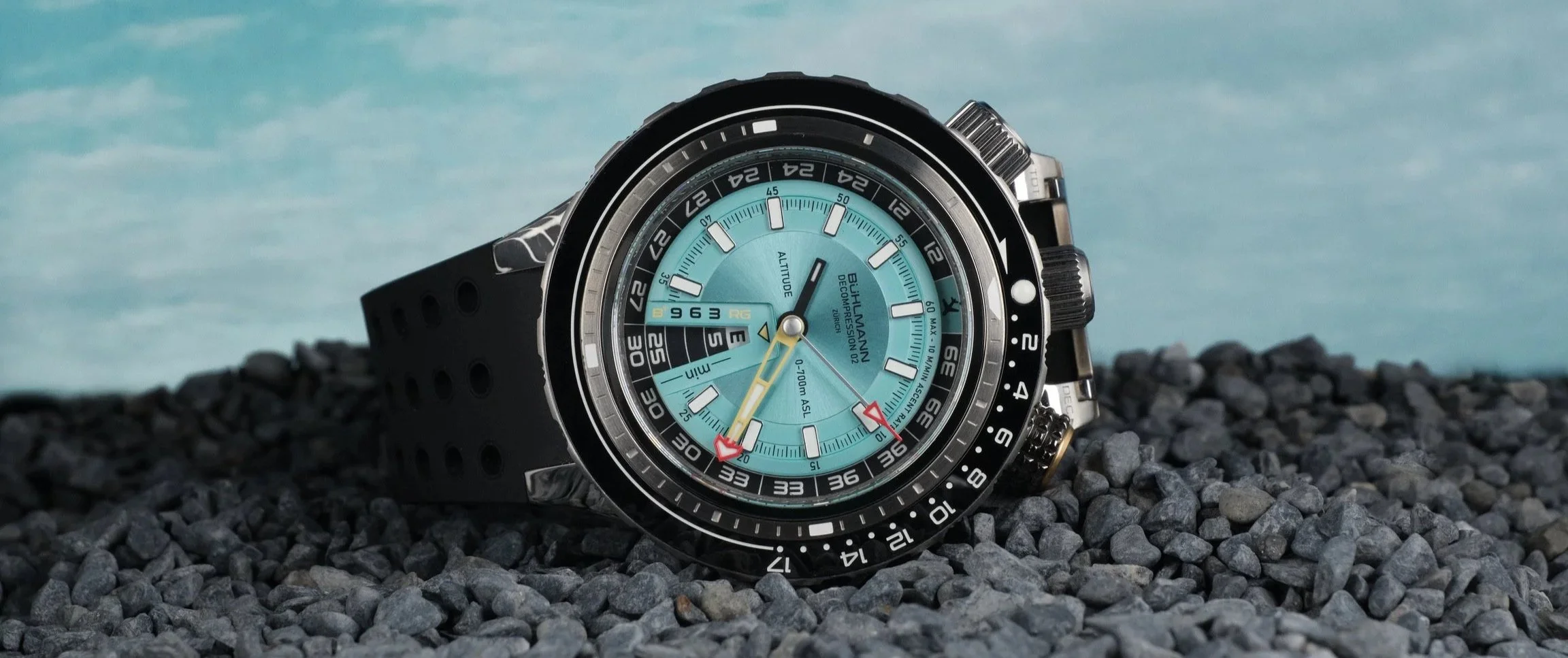 Buhlmann Decompression 02 dive watch with a turquoise decompression dial, multi-scale bezel, and oversized crown system, photographed on a black rubber strap against a rocky foreground and soft blue sky backdrop.
