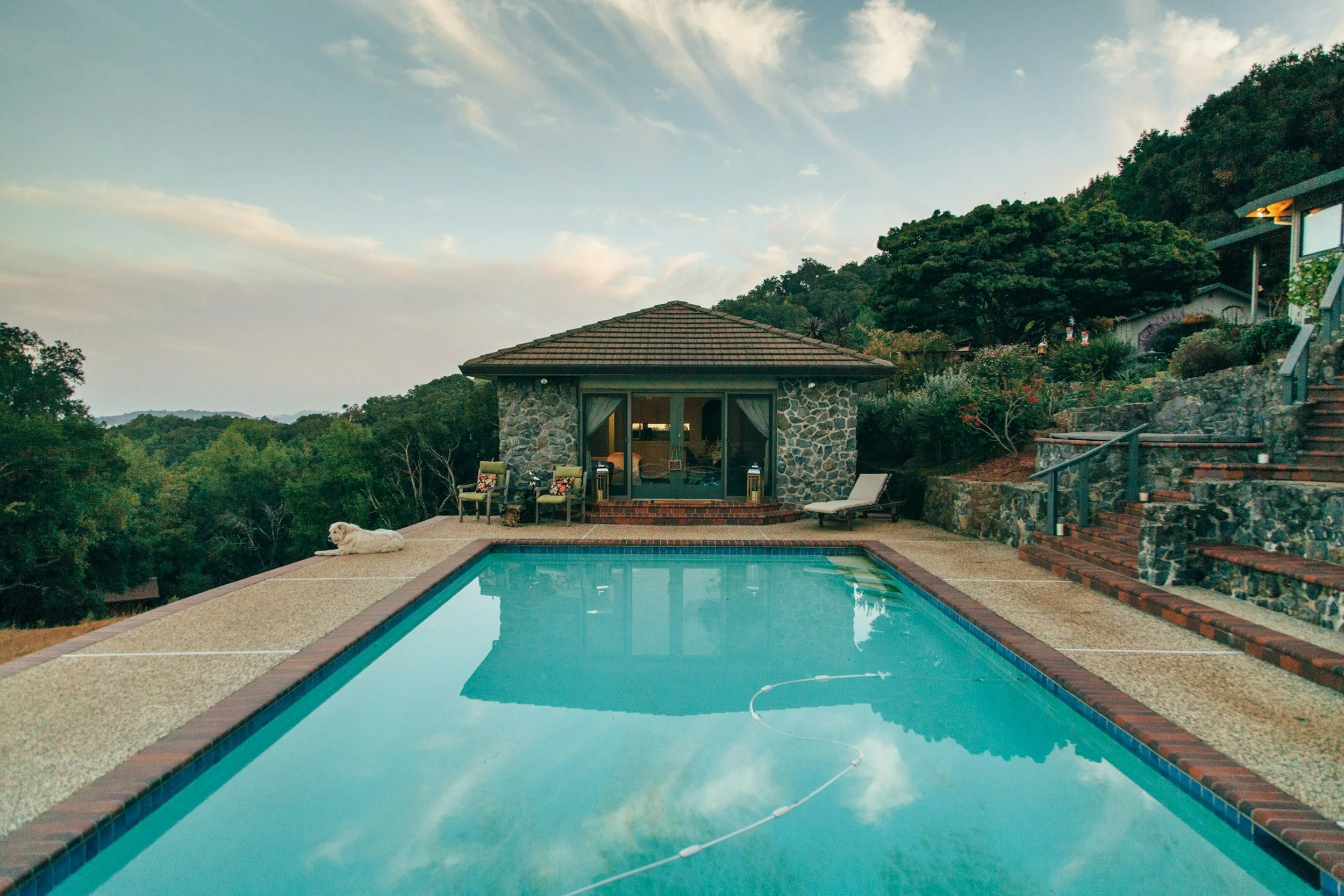 A backyard view of a swimming pool with calm water, reflecting the sky, surrounded by a paved deck, and a small stone-colored house or cabana with a tiled roof in the background. There is a white dog lying on the deck near the pool, and outdoor furniture including chairs and a lounge chair are visible. The scene is set in a scenic, hilly landscape with trees and greenery.
