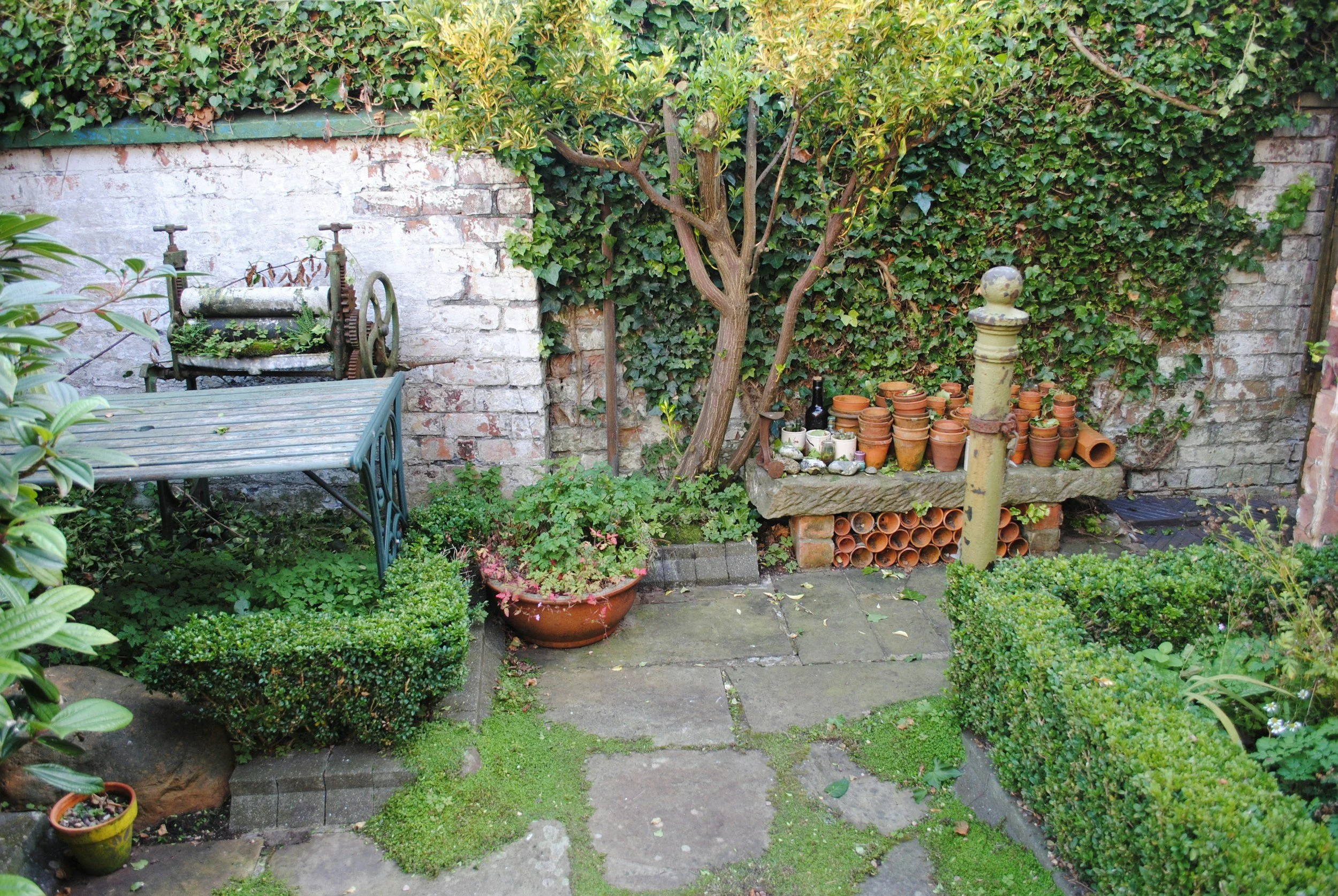 A cozy garden corner with potted plants, a vintage apple press, a metal table, a stack of clay pipes, and a small tree against a brick wall covered in ivy.
