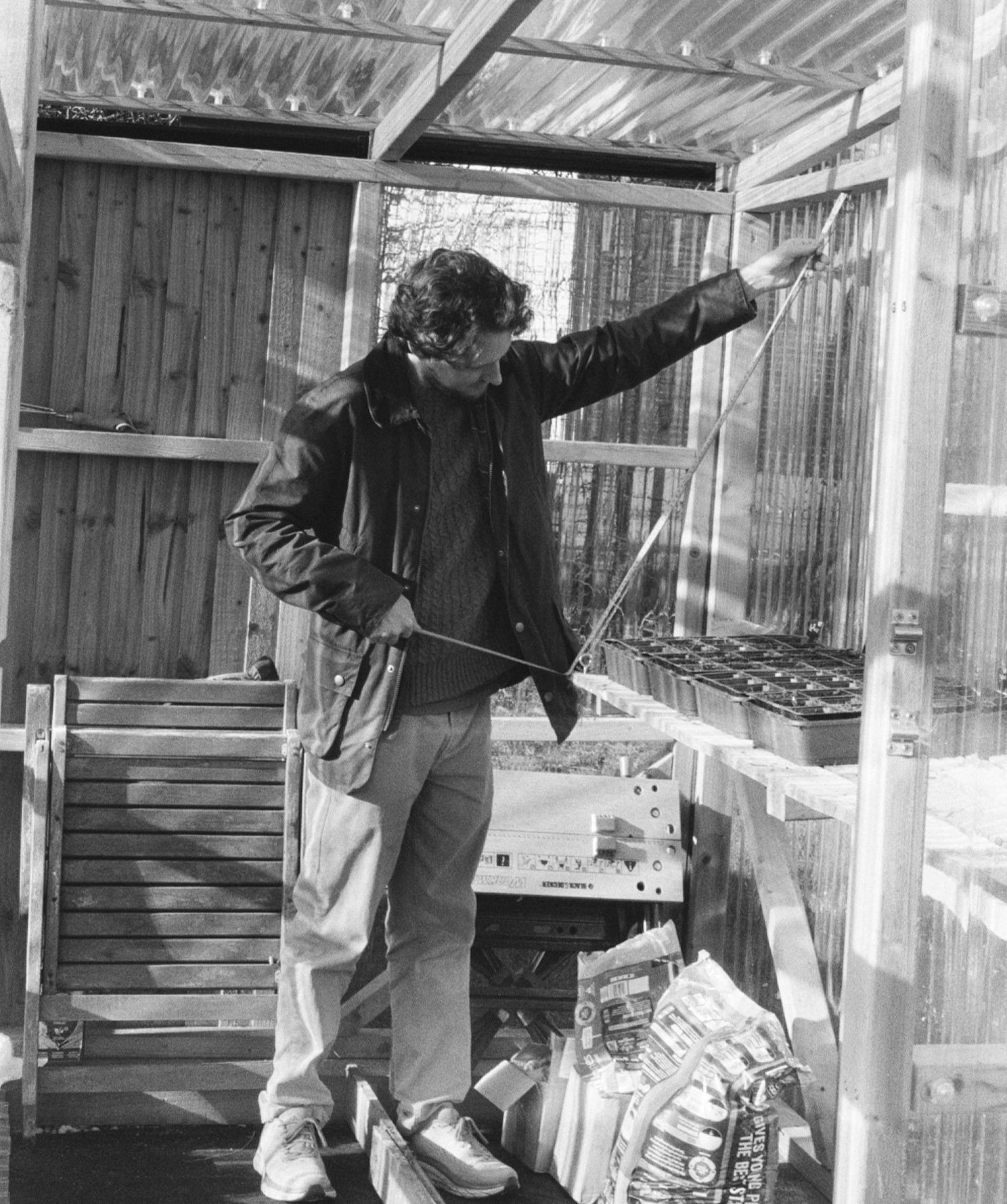 A person measuring the inside of a greenhouse with a tape measure, surrounded by gardening supplies and pots.