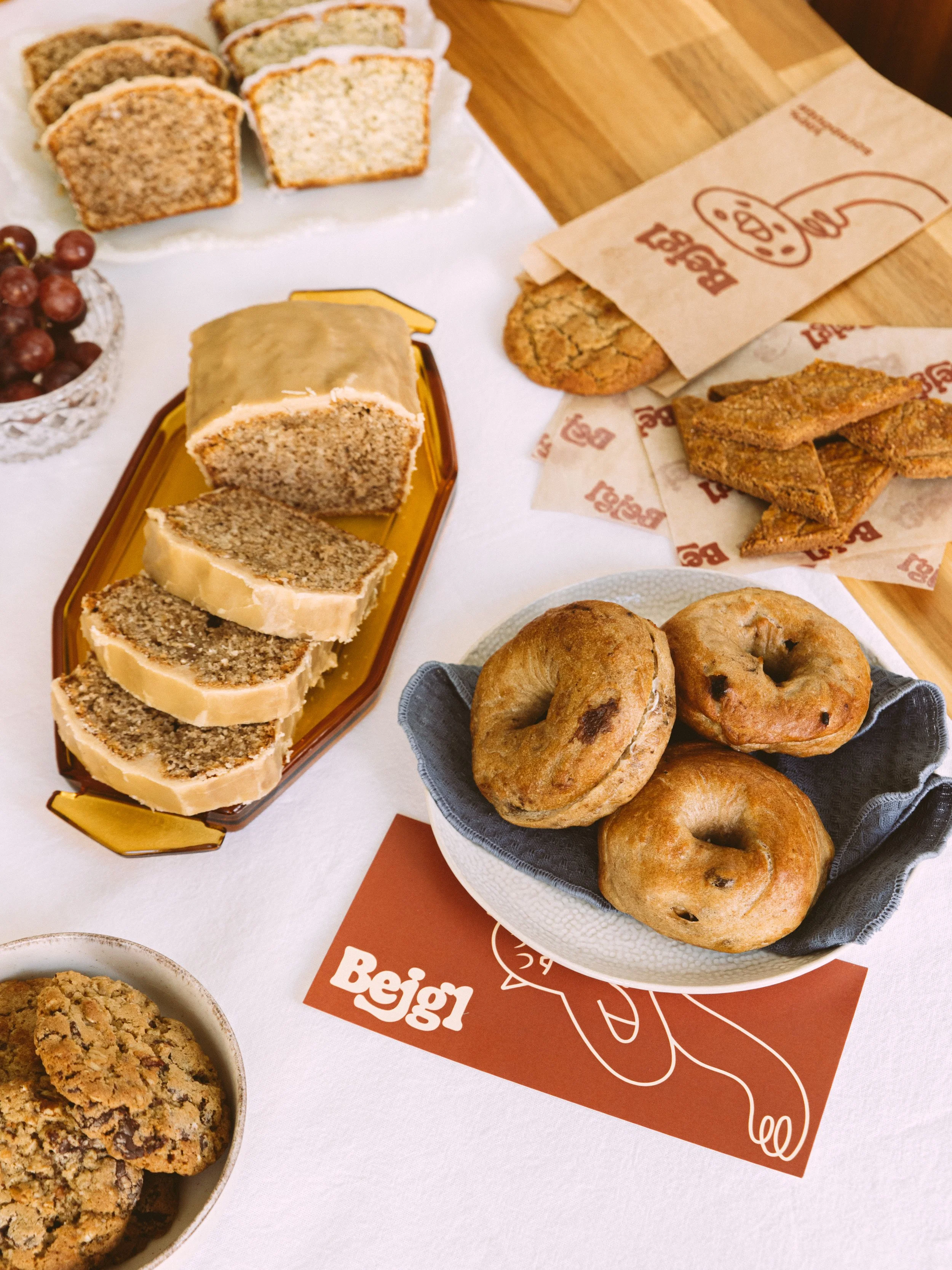 assortment of baked goods including sliced banana cake, cookies, bagels, breaded chicken, and pastries, arranged on a table with a white tablecloth and party-themed decorations.