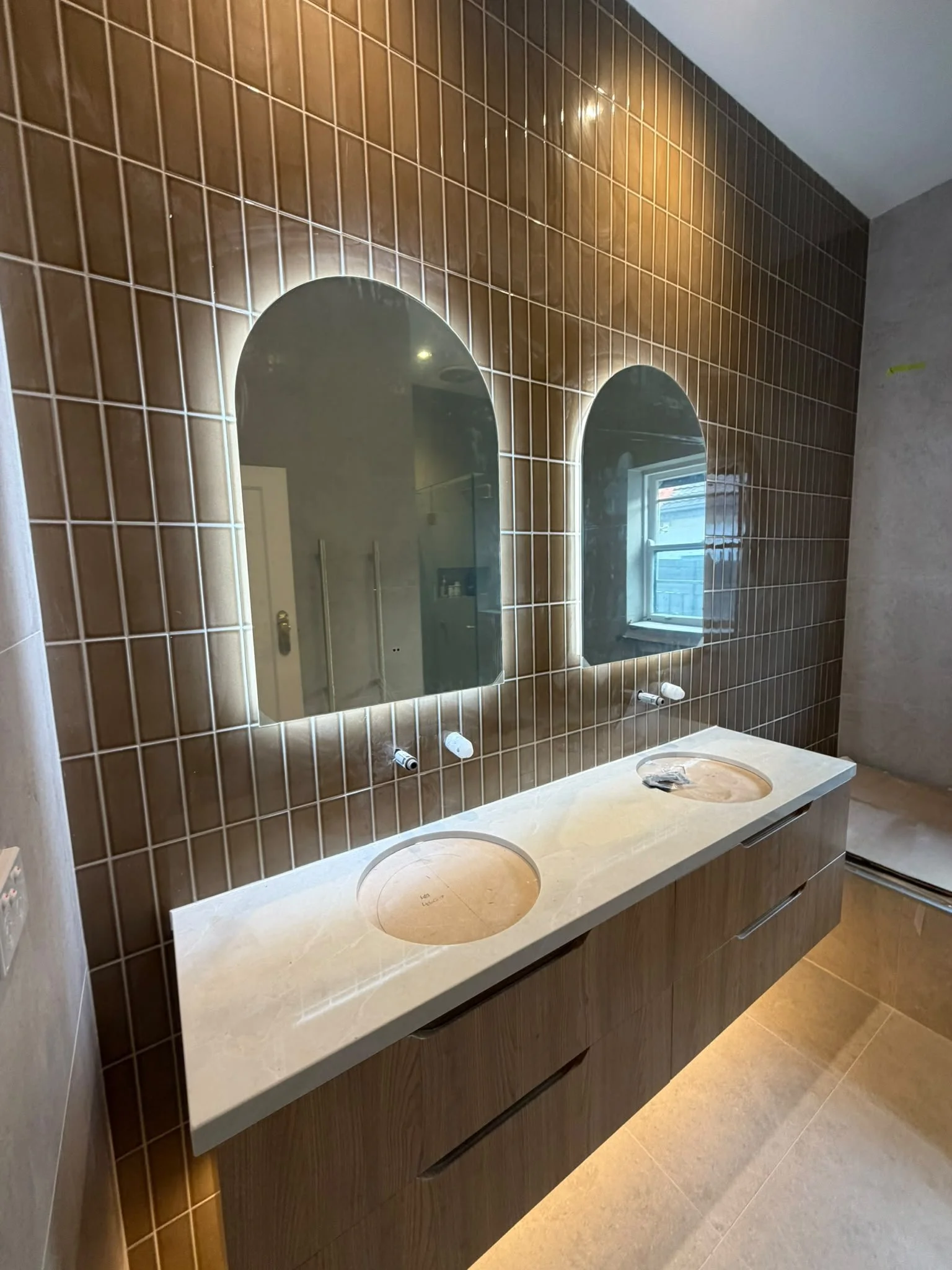 A modern bathroom vanity with a double sink, beige countertop, and two oval mirrors against a brown tiled wall, with window and lighting reflections visible.