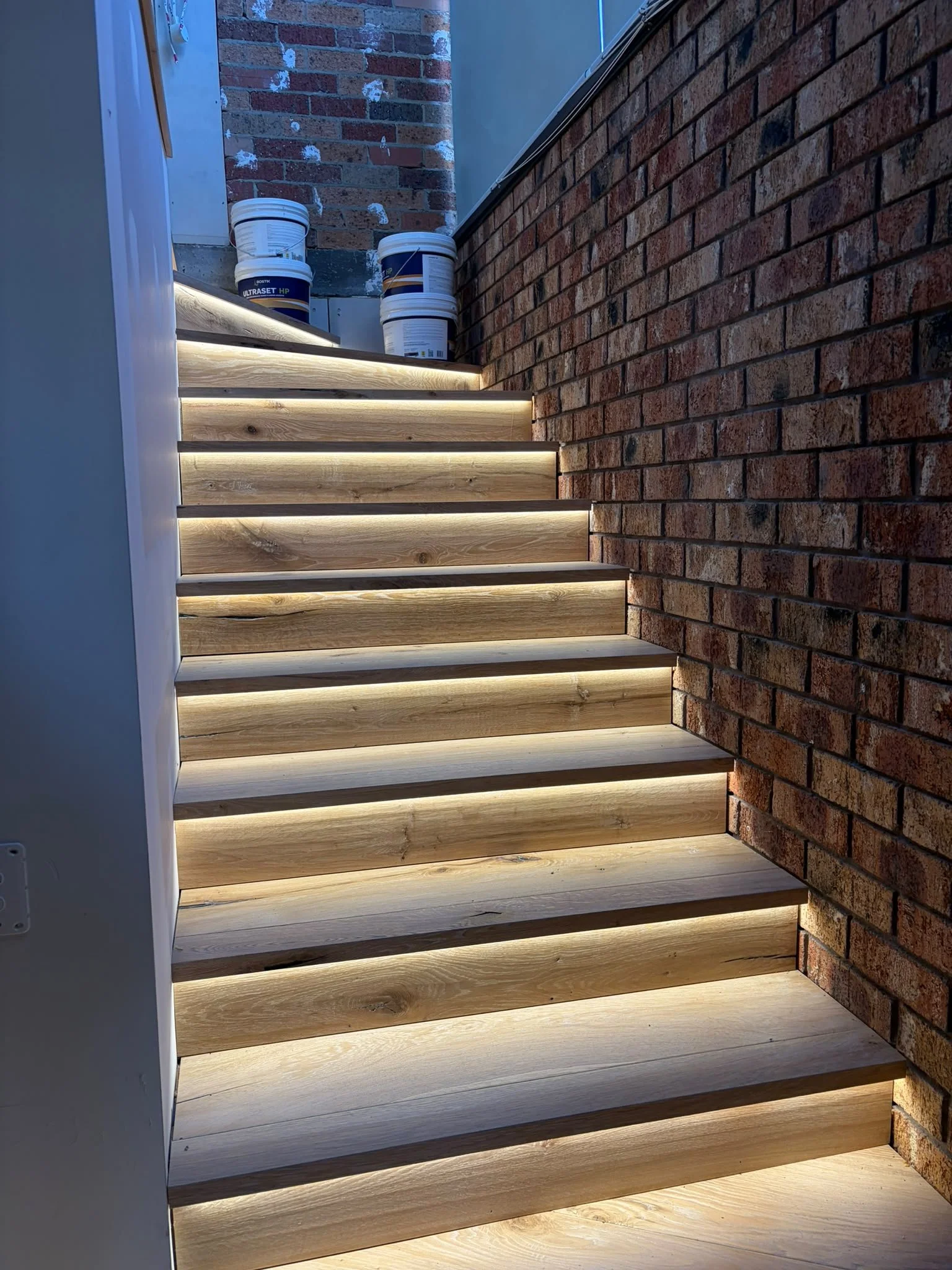 Wooden stairs with integrated LED lighting along the treads, set against a brick wall and a brick flooring background, with buckets and construction materials nearby.