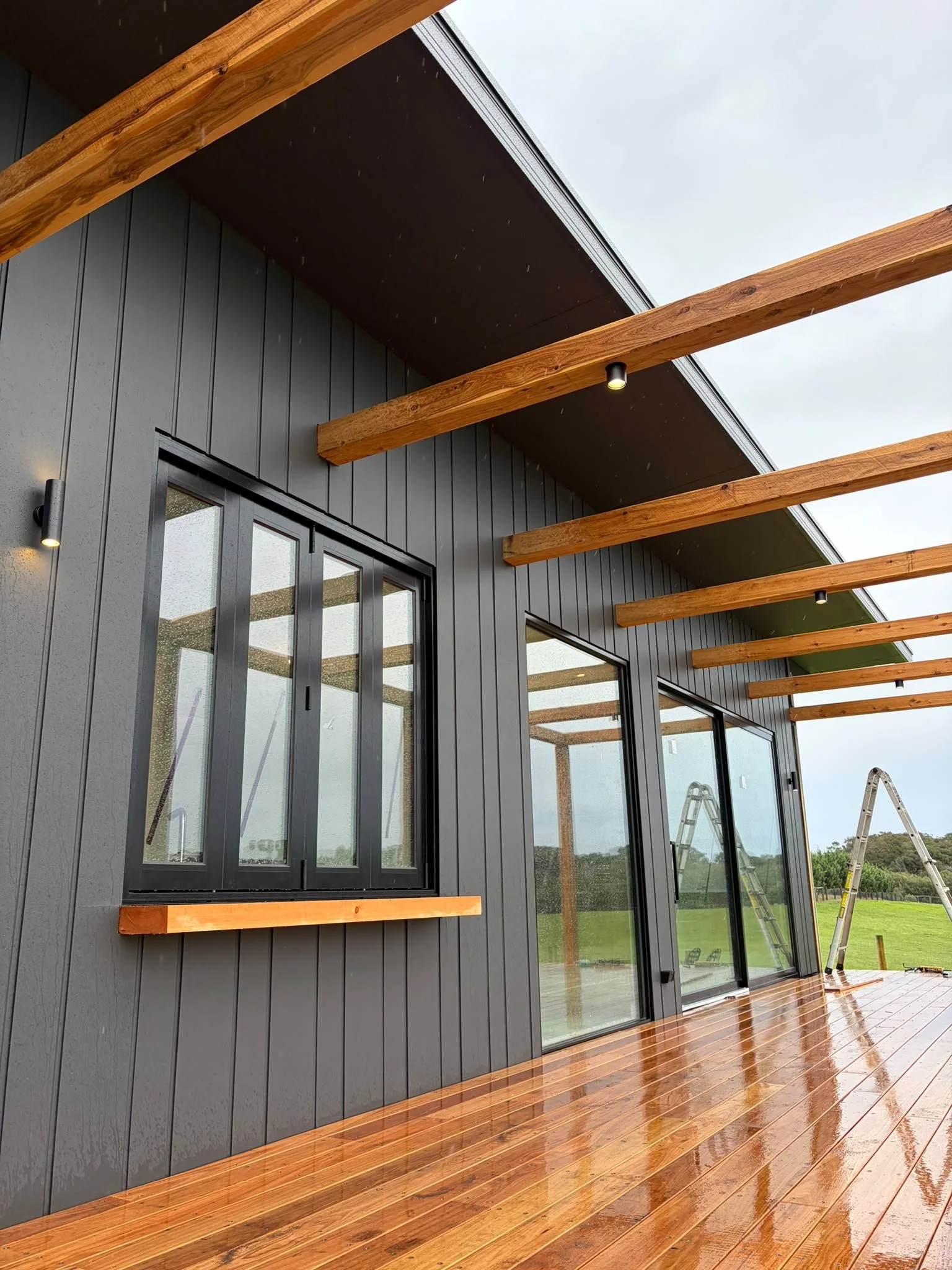 A modern house exterior with dark grey vertical siding, large glass sliding doors, wooden beams overhead, and a polished wooden deck, during rainy weather.