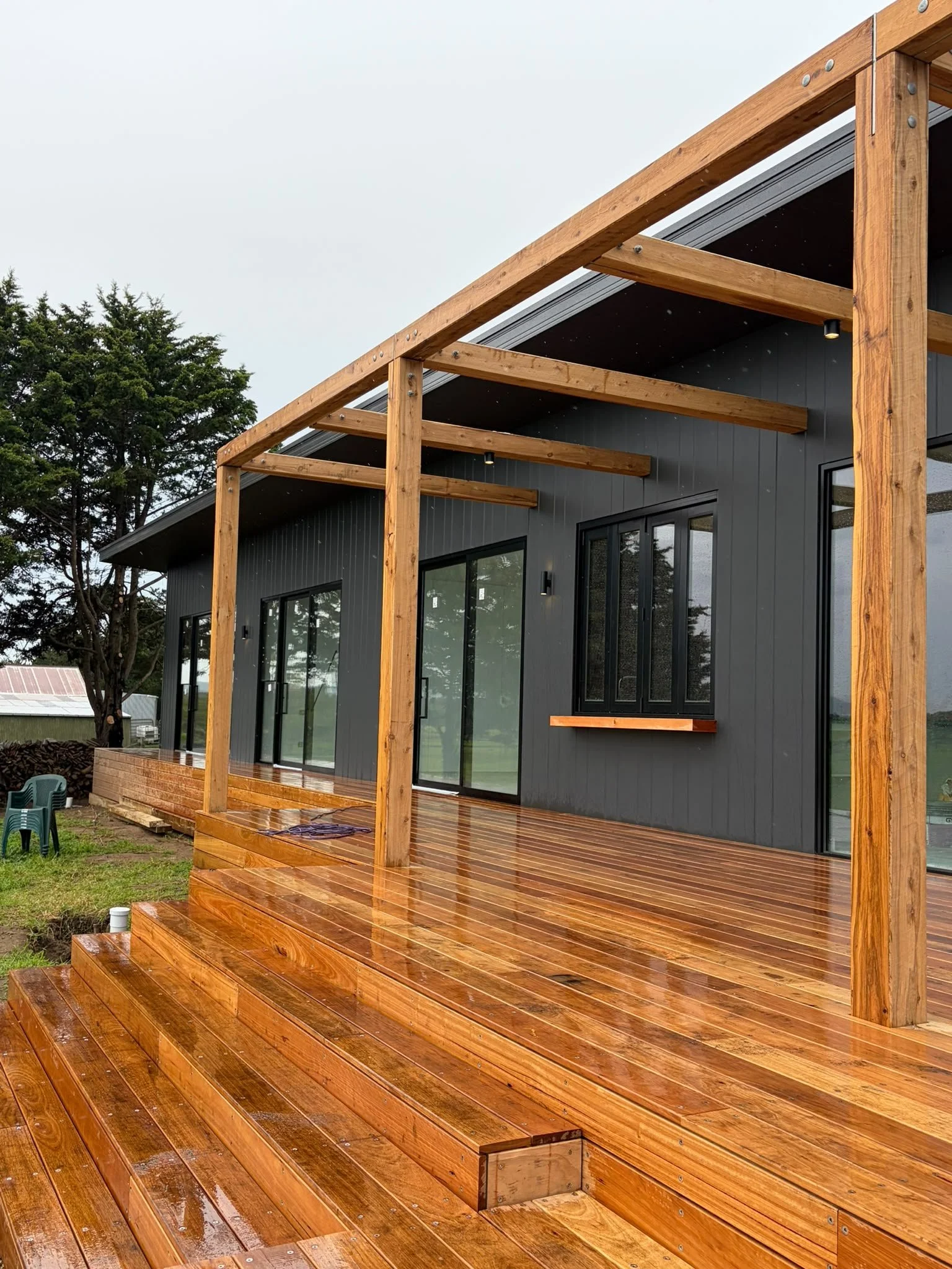 Wooden deck with steps leading to a modern house with large glass windows and dark gray siding. A wooden pergola frame is partially built over the deck. Trees and a cloudy sky are in the background.