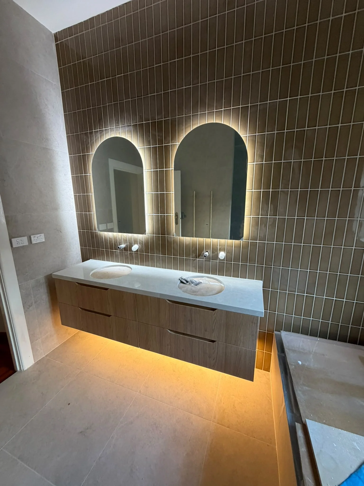 Bathroom vanity with two oval sinks, large mirror with LED backlighting, beige tile walls and floor, electrical outlets on the wall, and a marble countertop.