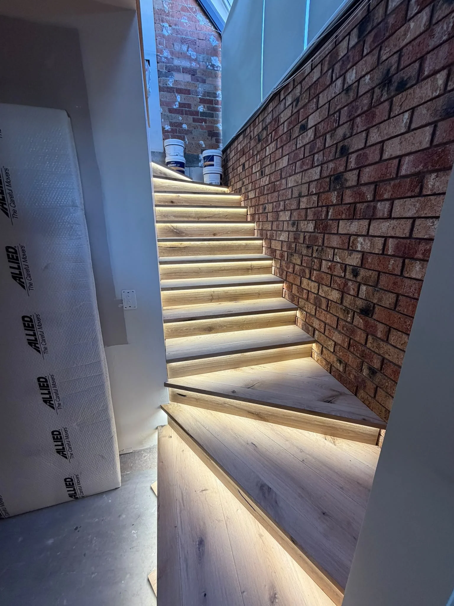Interior of a building under construction showing exposed brick walls, wooden stairs with lighting, and construction materials like buckets.