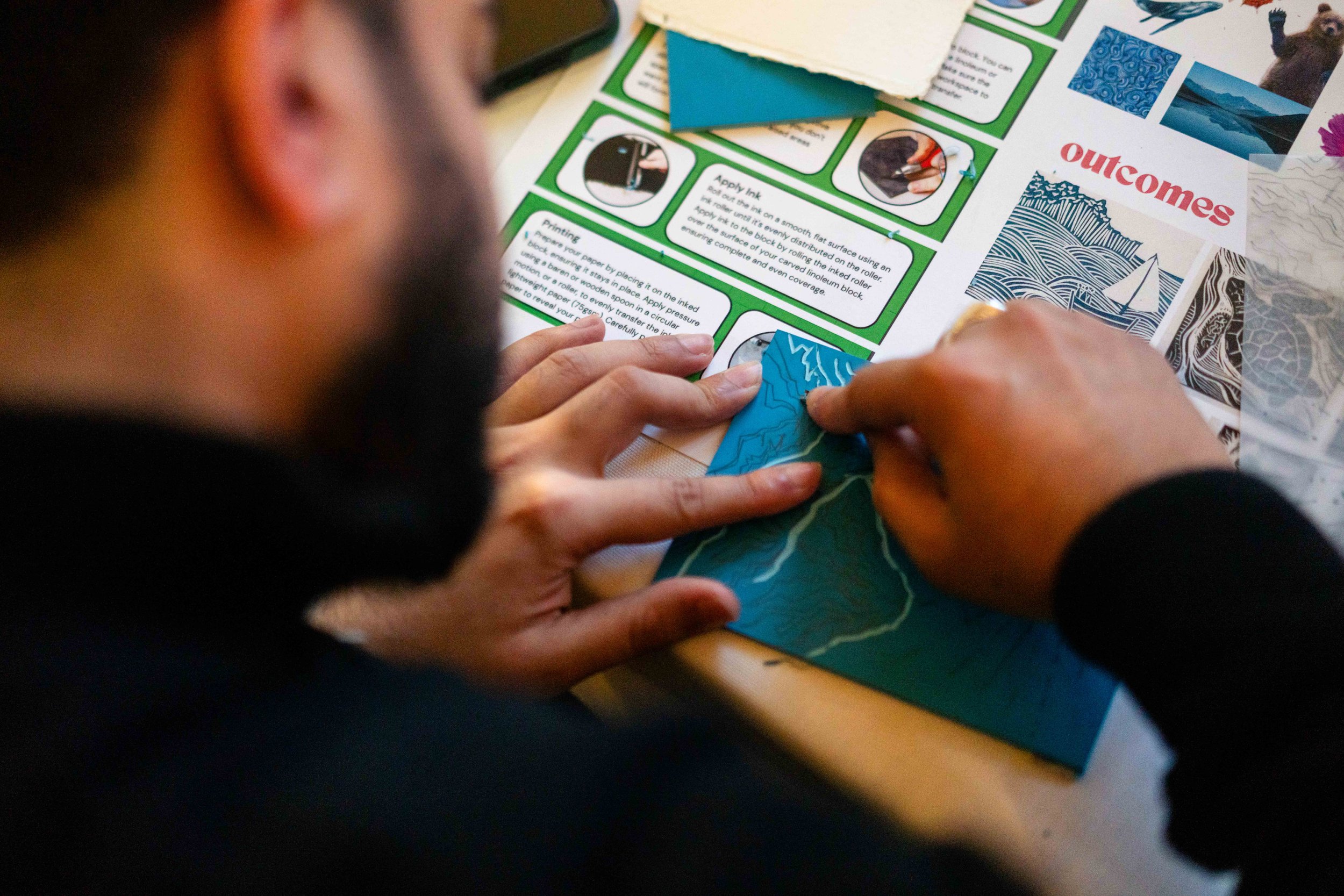 Person carving a design into a blue linoleum block, surrounded by art tools and printed instructional materials.