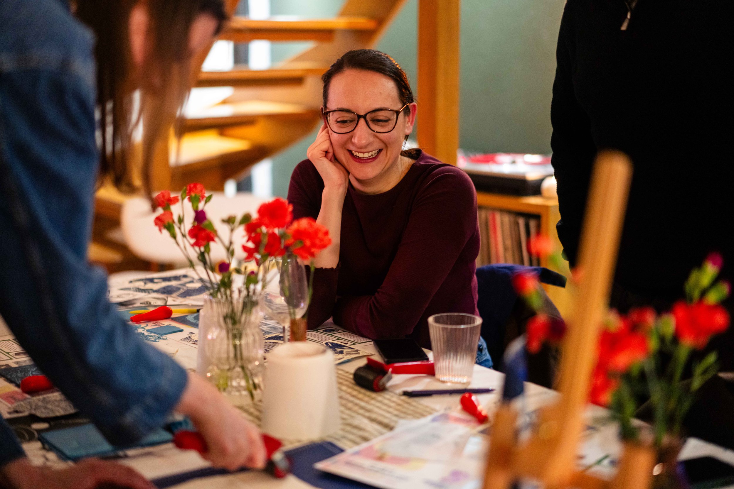 Group of people engaging in arts and crafts at a table with red flowers in vases and art supplies.
