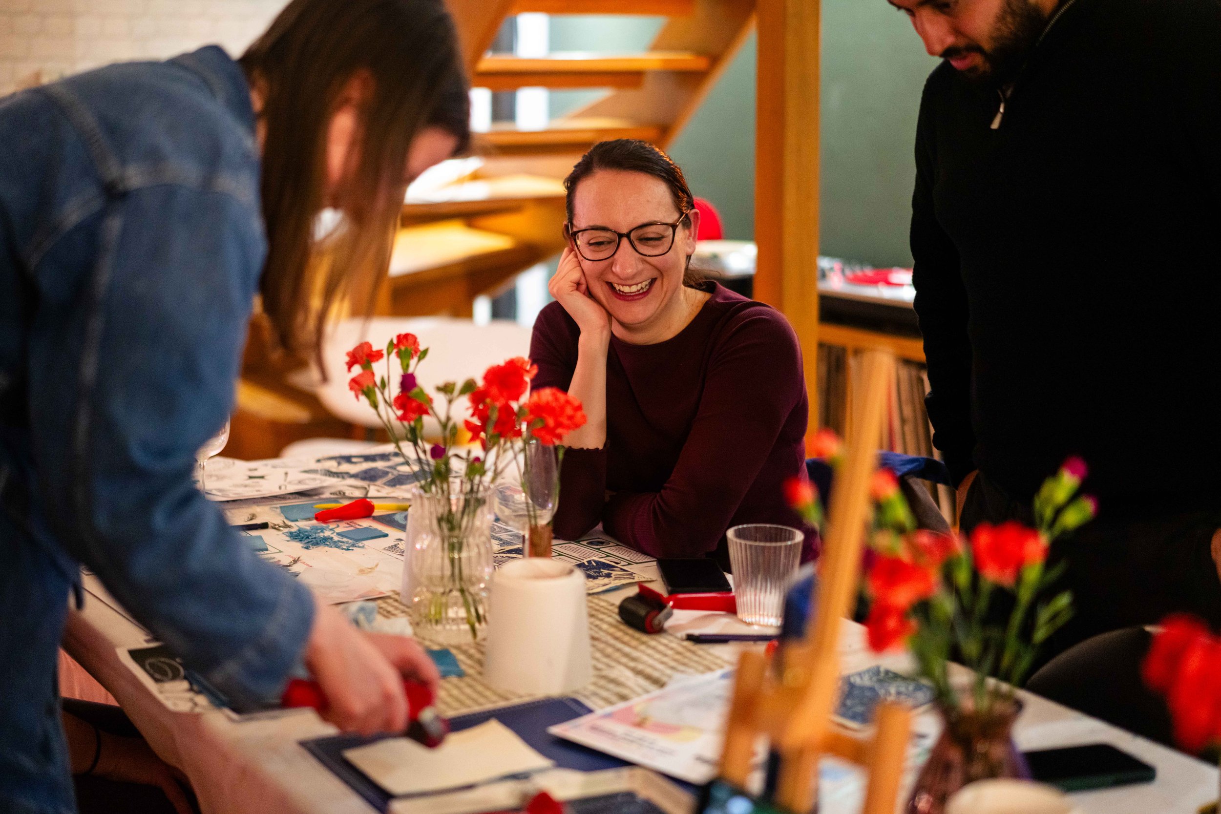 Three people engaged in an arts and crafts activity at a table decorated with flowers, art supplies, and papers. One person is using a roller tool, while another smiles while watching. The setting is indoors with a cozy atmosphere.