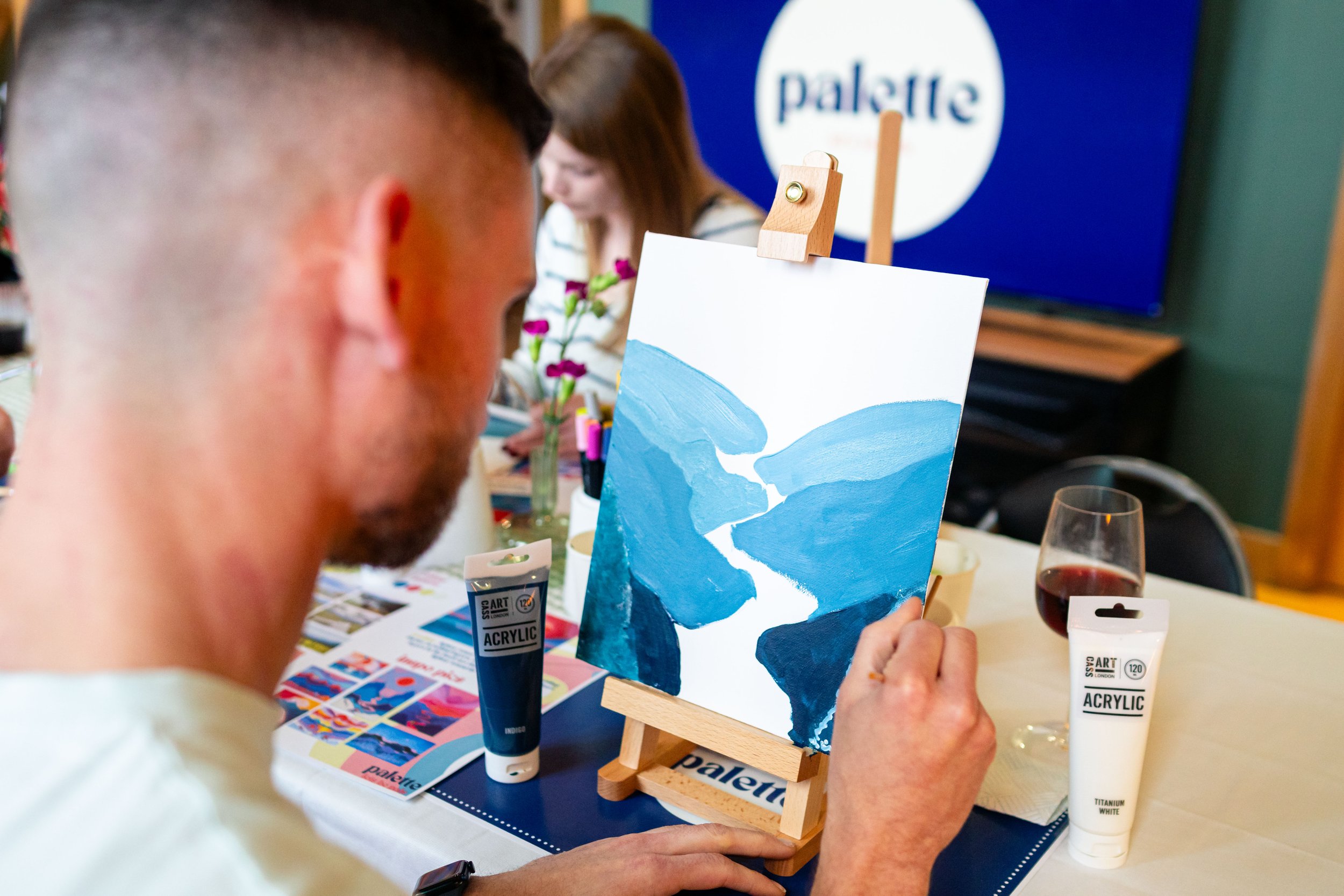 Person painting on canvas with blue acrylic paint, glass of red wine on table, art supplies, and event setting.