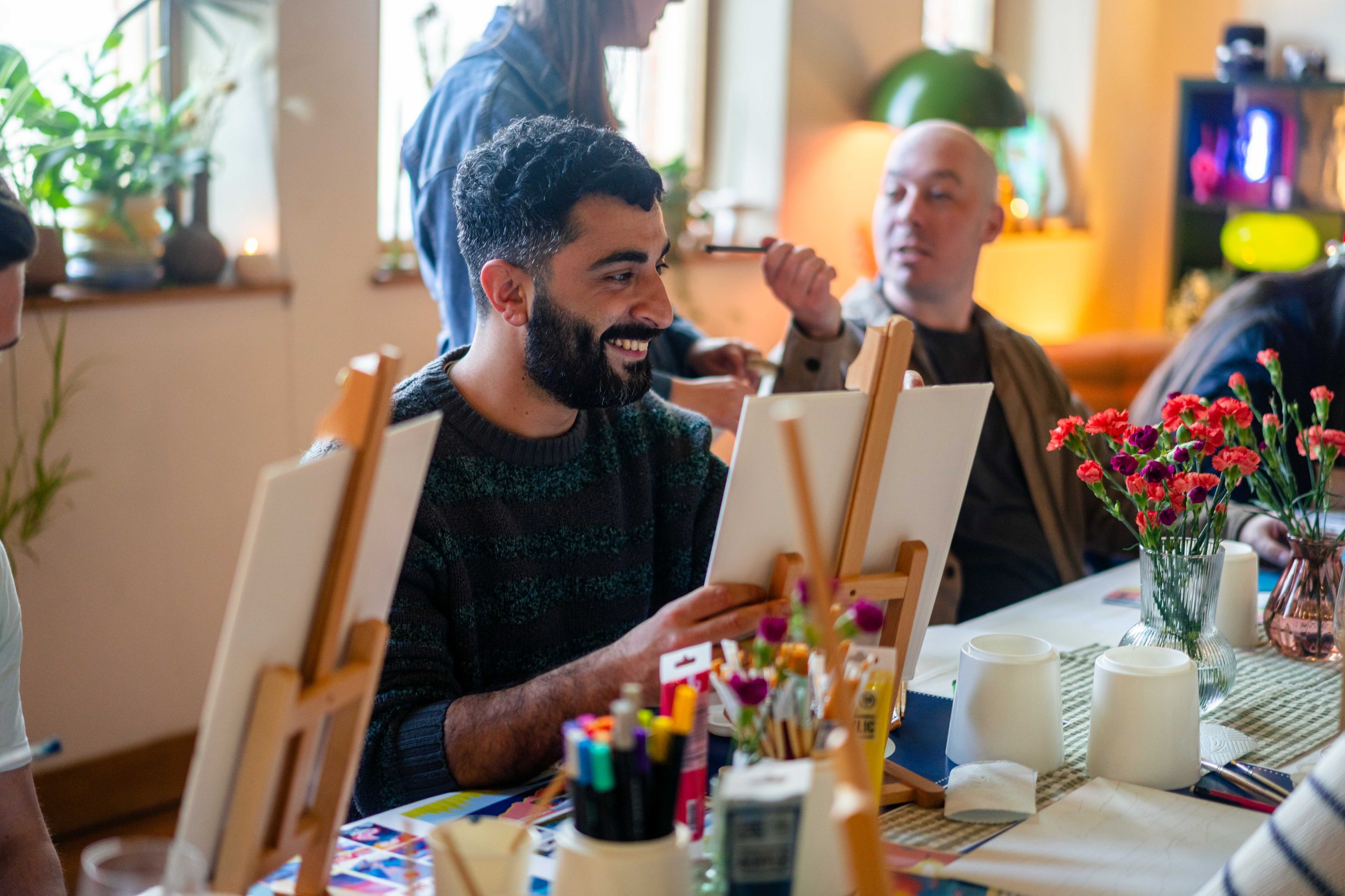 People sitting around a table engaged in a painting activity with easels, surrounded by art supplies and vases of flowers in a well-lit room with plants.