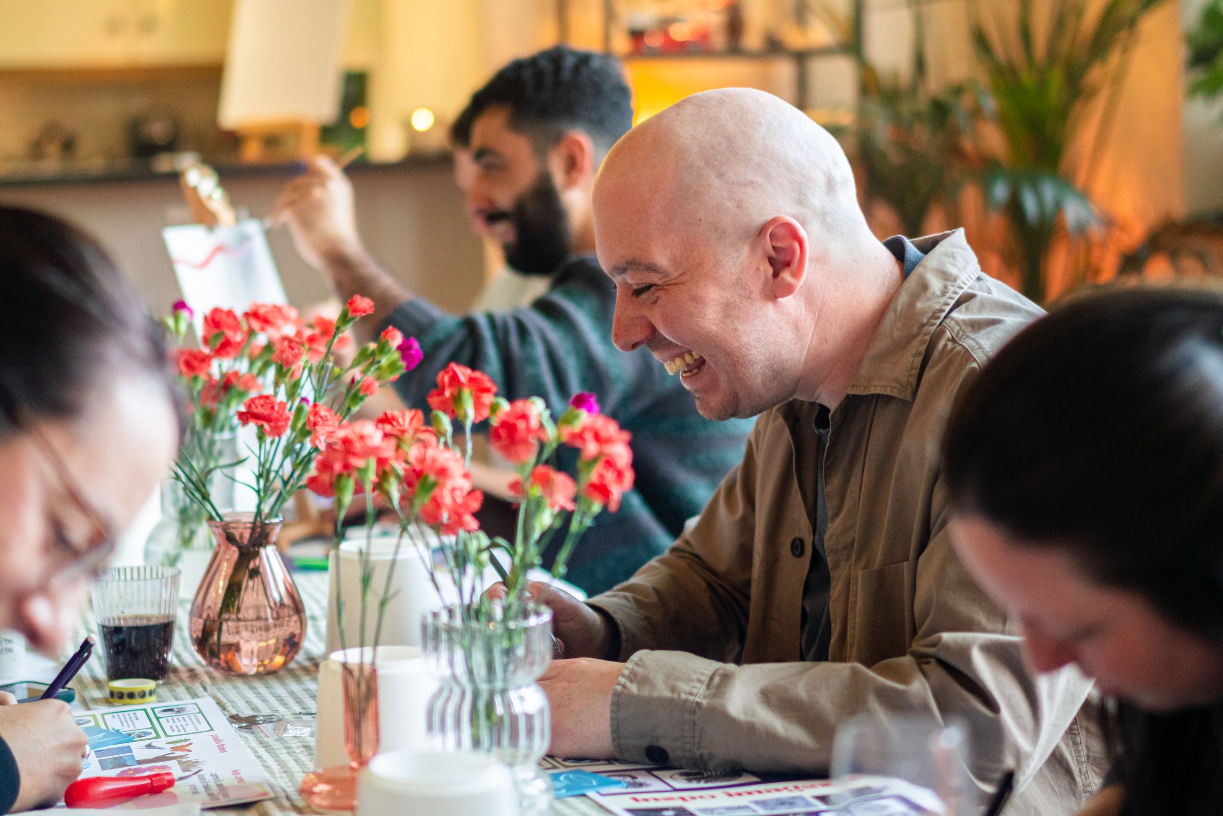 People gathered around a table, smiling and engaged in an art activity. Red carnations in vases are on the table.