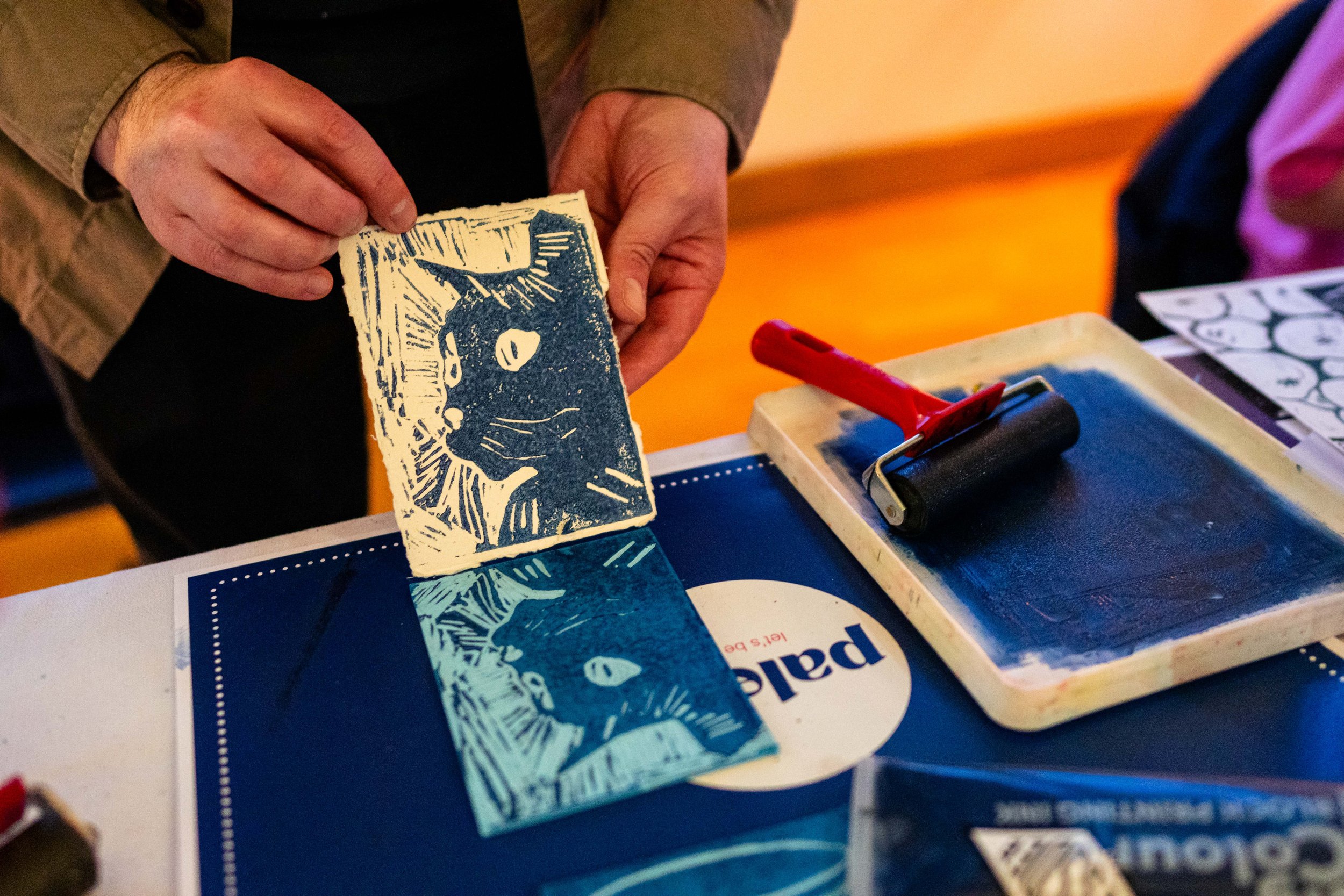 Person holding a linocut print of a cat next to a roller and ink setup on a table.