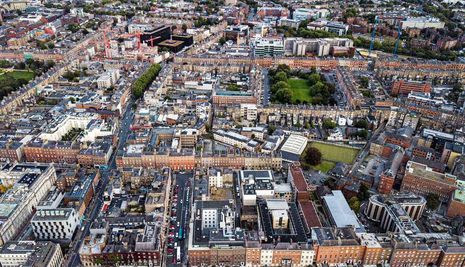 Aerial view of a dense cityscape featuring a mix of modern and historic buildings, with some construction cranes, green spaces, and streets lined with parked cars.