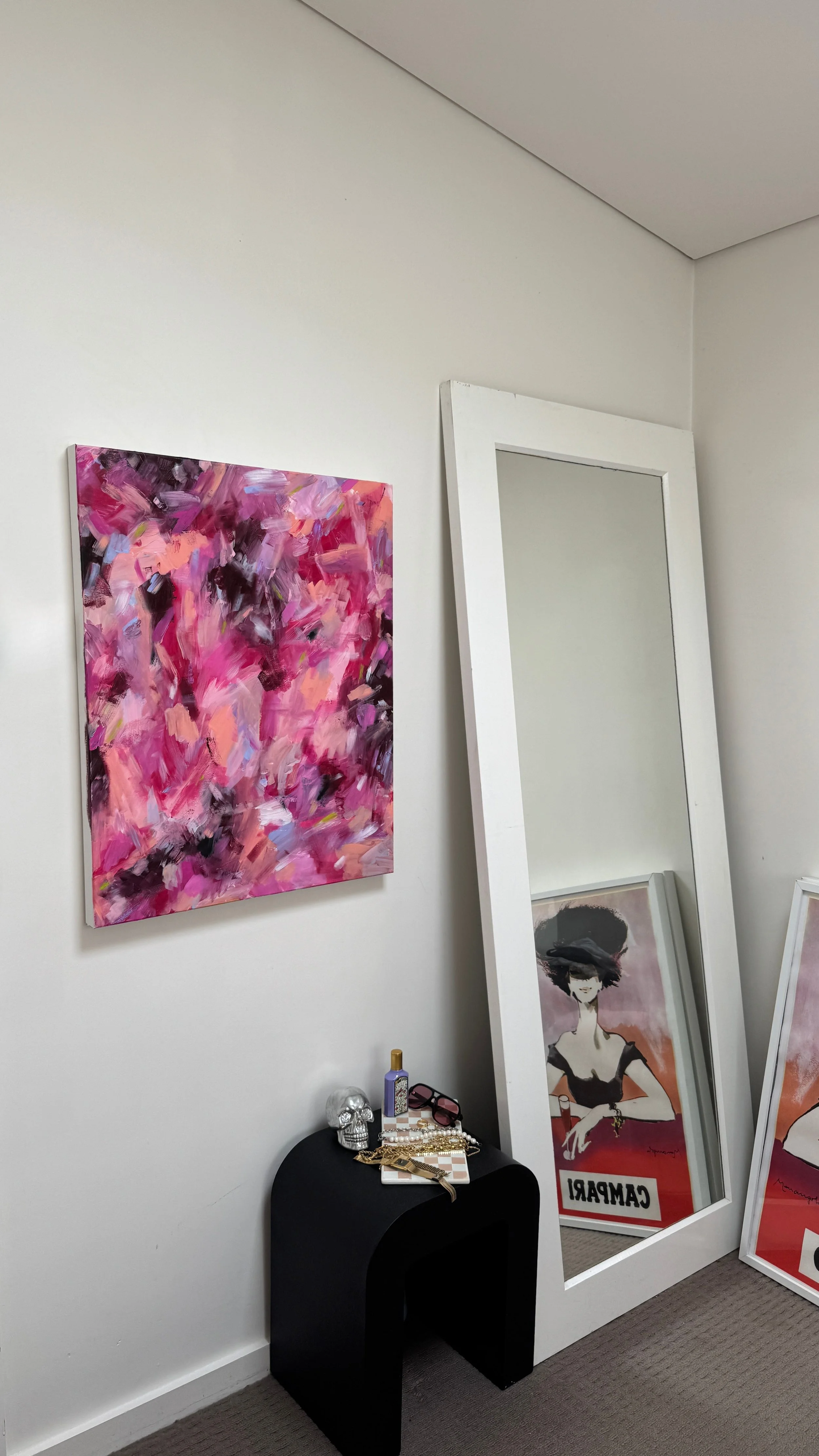 A colorful abstract pink and purple painting on a white wall, a large white-framed mirror leaning against the wall, and framed fashion illustrations with one labeled "CAMPAIGN" on the floor. Small decorative items are on a black table below the painting.