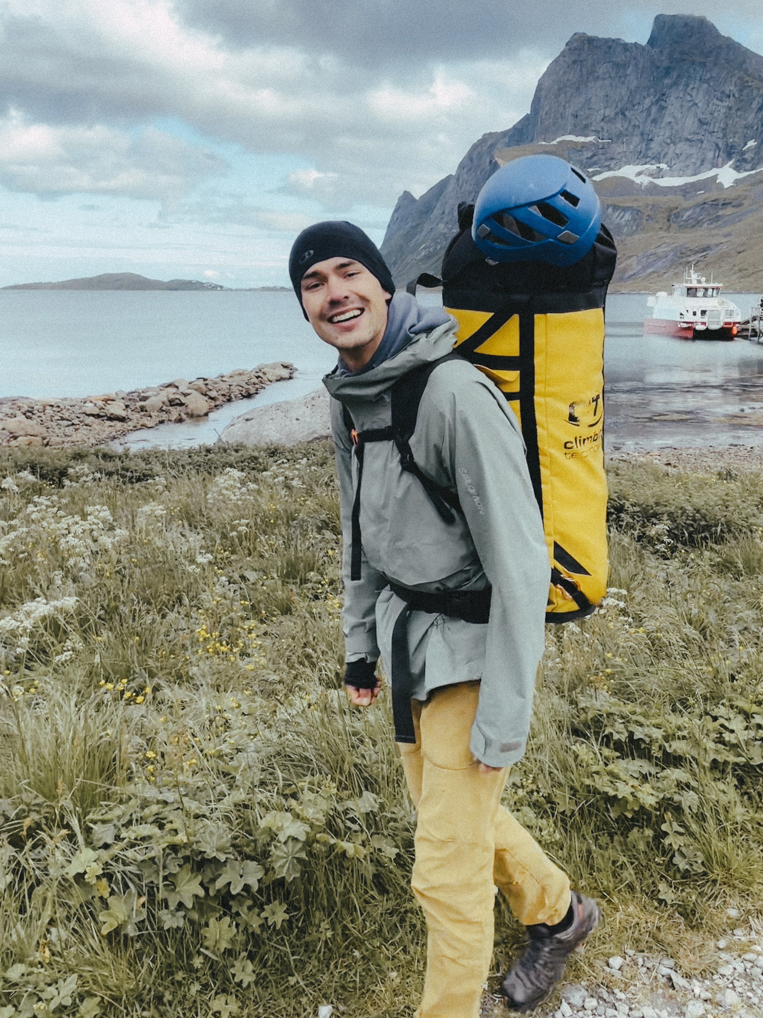 Junger Mann beim Wandern an einem See mit Bergen im Hintergrund, trägt eine Klettersicherung und einen Rucksack mit Helm