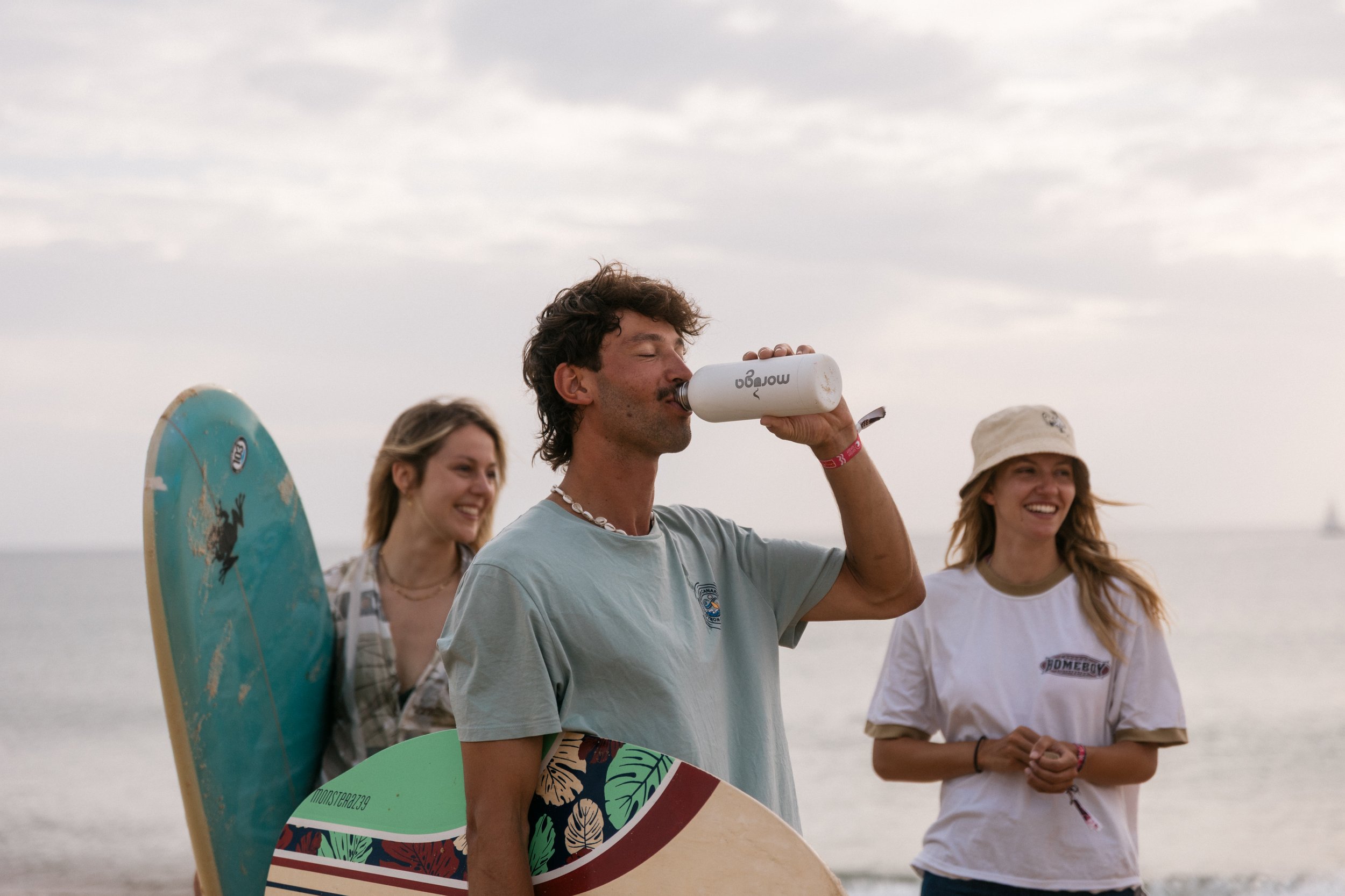 Drei junge Menschen am Strand, eine Frau hält ein Surfbrett, ein Mann trinkt aus einer Wasserflasche, alle lachen, im Hintergrund das Meer und ein Boot.