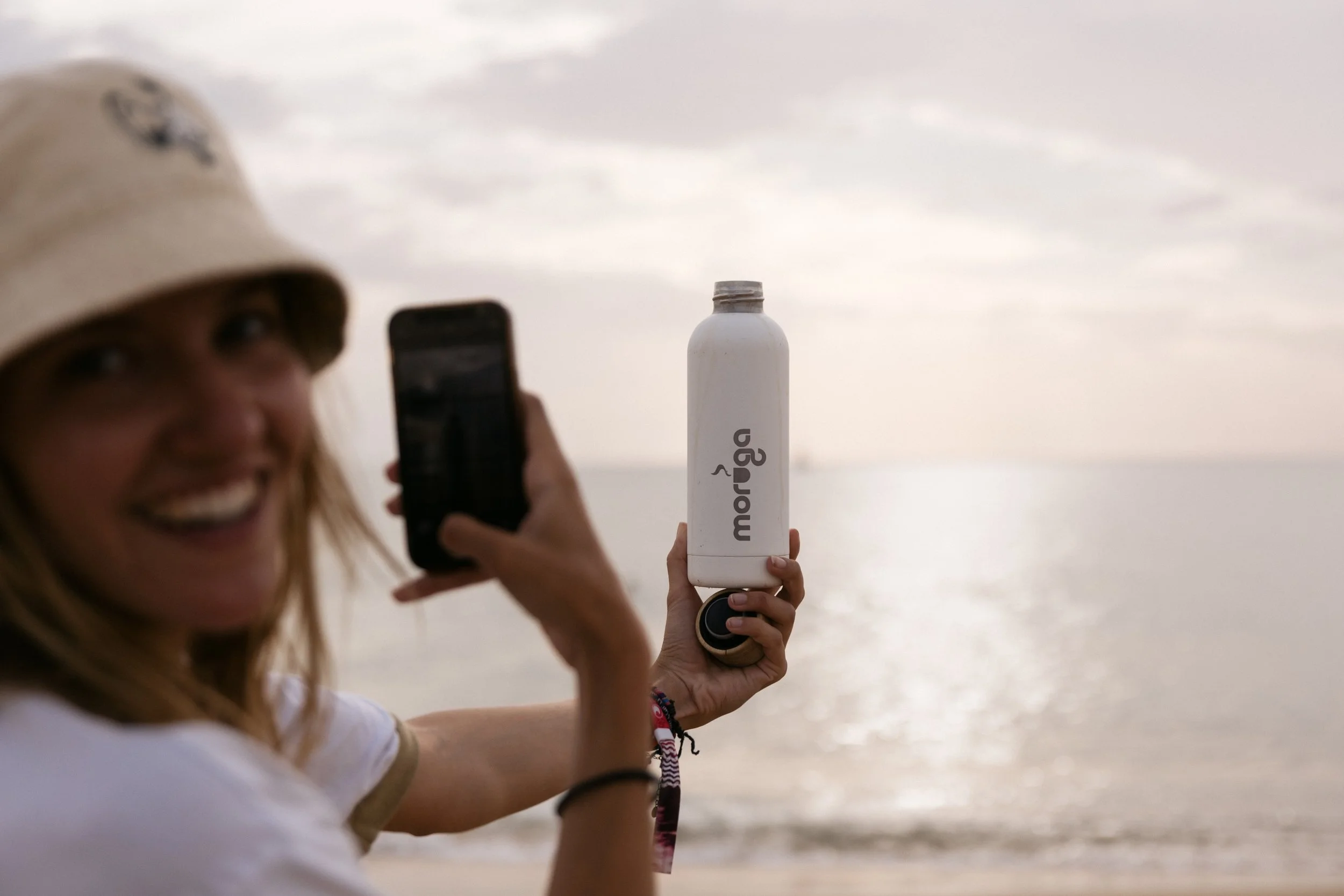 Frau hält eine weiße Wasserflasche mit dem Markennamen 'marég' vor einem Strand bei Sonnenuntergang, macht ein Selfie.