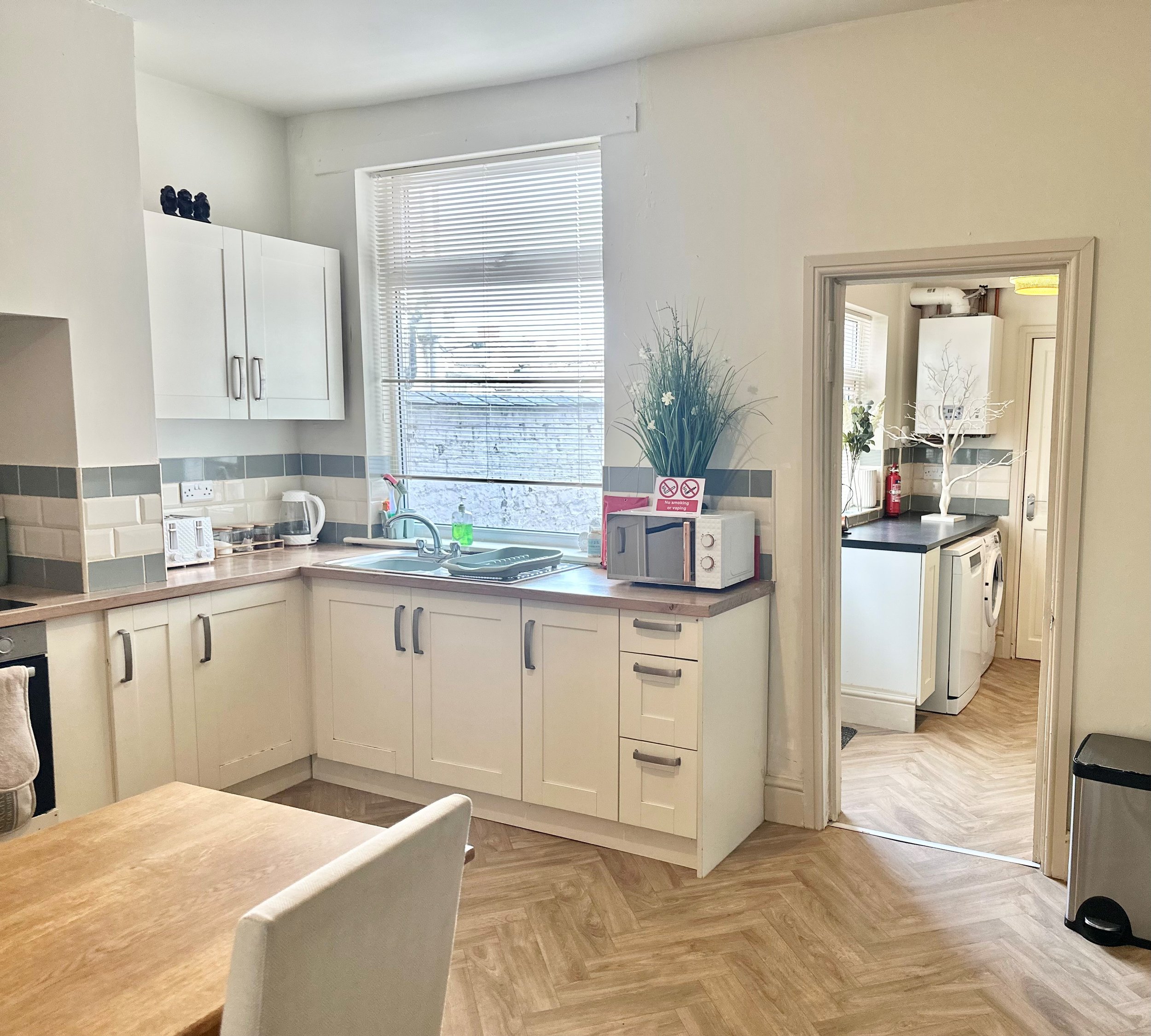 A kitchen with white cabinets, a window with blinds, beige countertops, and various appliances including a microwave, kettle, and toaster. There is a vase with tall artificial grass and a microwave on the countertop. An open doorway leads to a laundry area with a washing machine and dryer, and a small table or cabinet with a decorative tree.