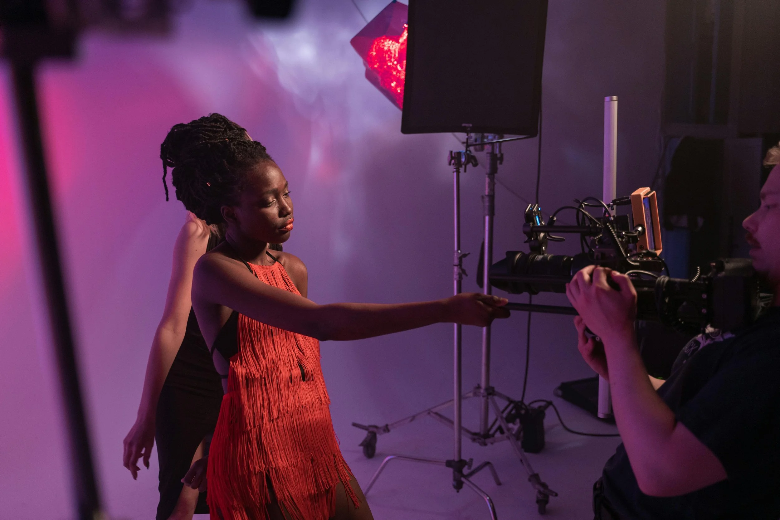 Una donna con abito rosso che fa una posa in uno studio fotografico, mentre un operatore con videocamera la riprende.