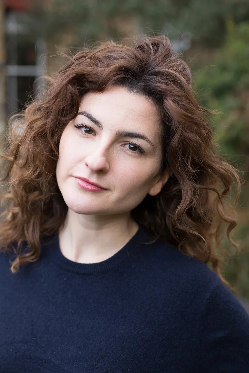 A woman with curly brown hair wearing a navy blue top, looking at the camera with a slight smile, outdoors with blurred trees in the background.
