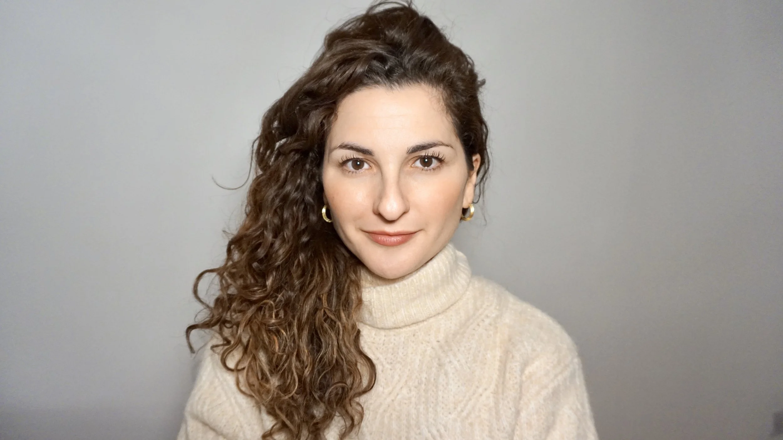 A woman with long, curly brown hair tied to one side, wearing a cream turtleneck sweater and gold hoop earrings, smiling softly at the camera against a plain gray background.