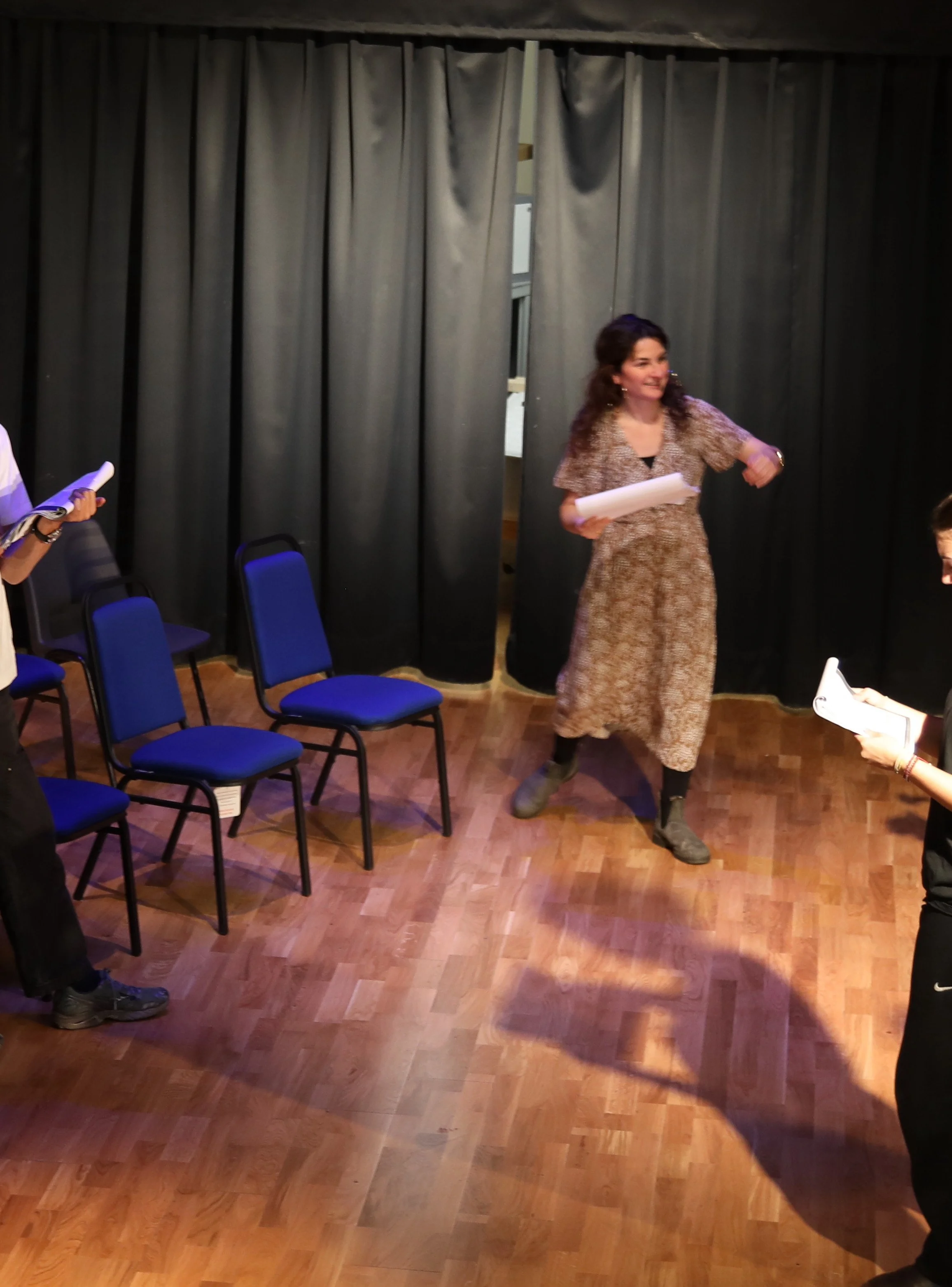 A woman with dark curly hair wearing a patterned dress and black boots standing on a wooden floor in front of dark curtains, holding sheets of paper, smiling, and gesturing with her right hand. Two other people holding papers are partially visible on the sides.