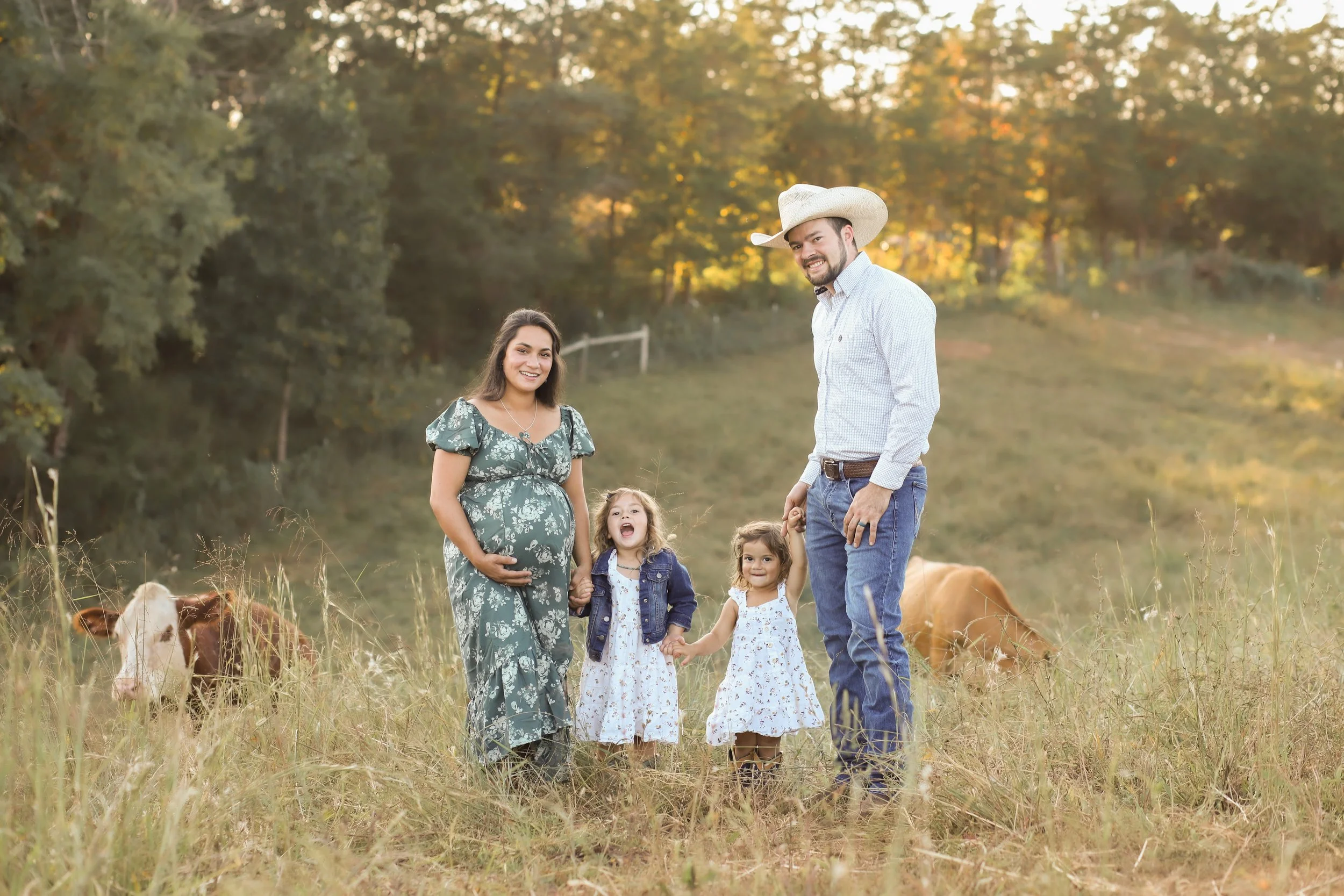 Family of five standing in a grassy field with cows, trees, and sunset in the background. Pregnant woman in floral dress, man in cowboy hat and jeans, three young girls in dresses and jackets.