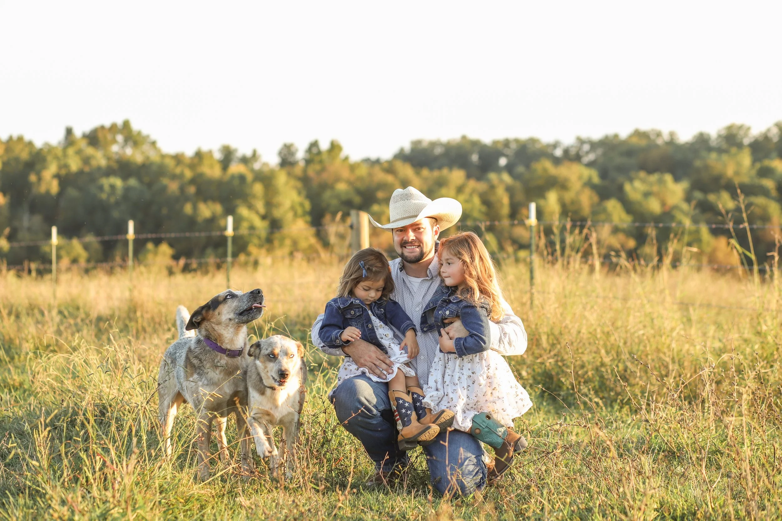 A man wearing a cowboy hat happily holding two young girls in a grassy field with three dogs, and a fence in the background at sunset.