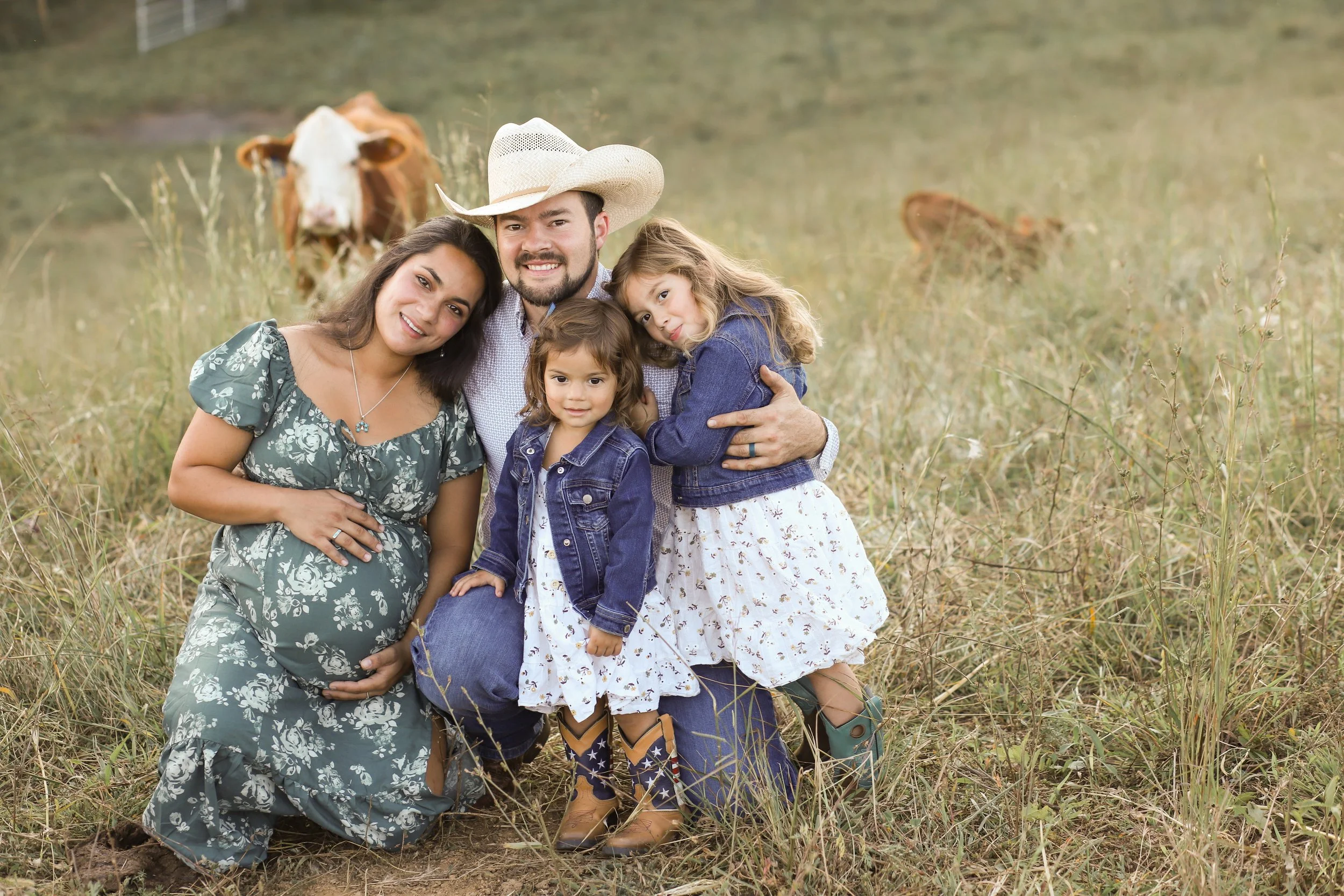 A family of five posing in a farm field with cows in the background. The family includes a pregnant woman in a floral dress, a man wearing a cowboy hat and plaid shirt, and three young girls dressed in denim jackets and dresses.
