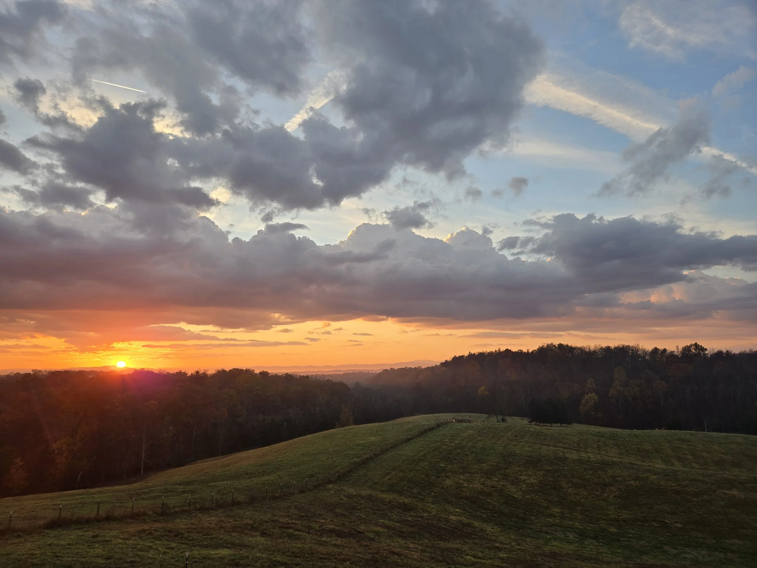 Sunset over rolling green hills with a tree line in the distance and a partly cloudy sky with vibrant orange and yellow hues.