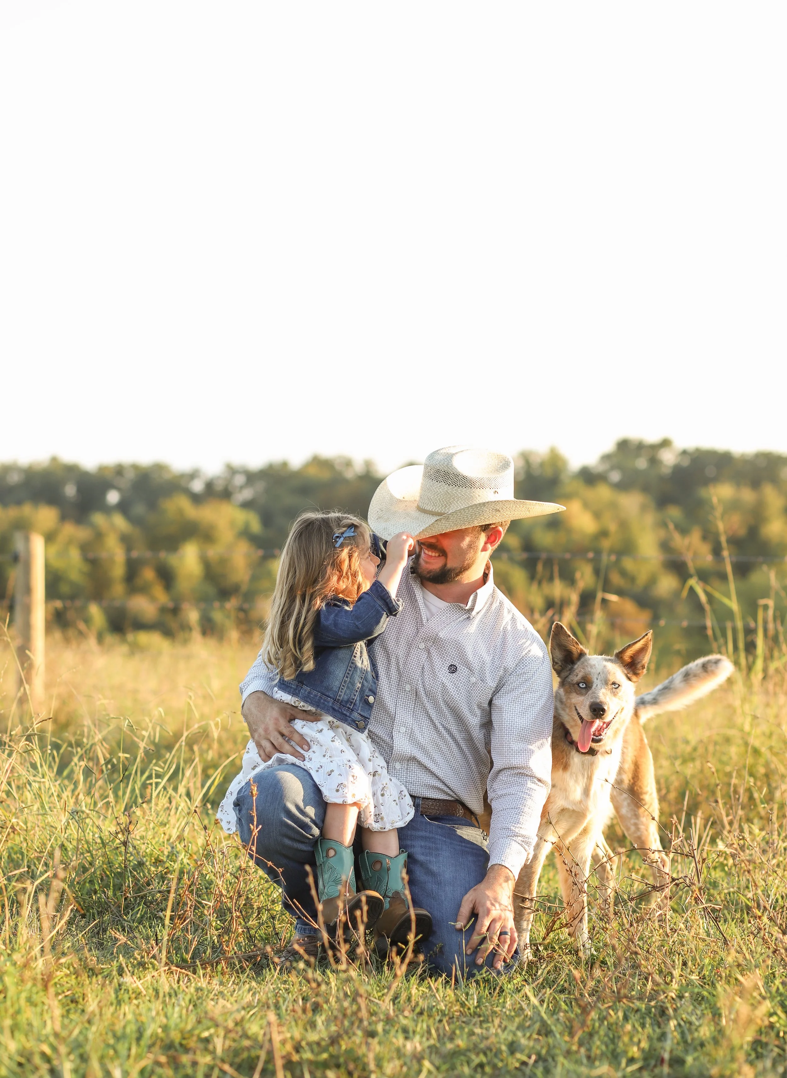 A man with a cowboy hat, kneeling in the grass, holding a young girl and a brown and white dog in a rural field during sunset.