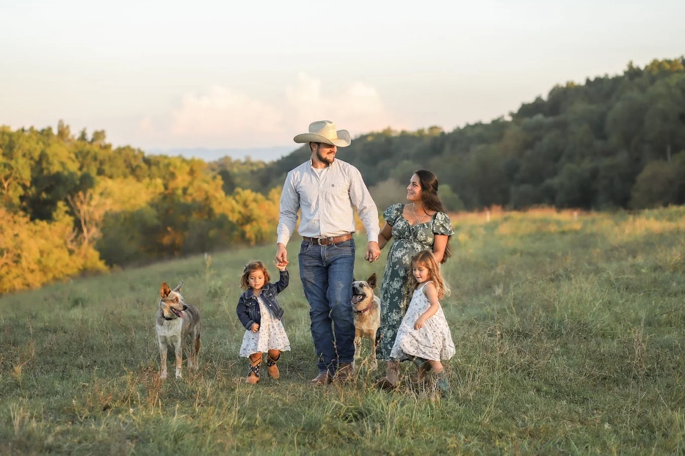 A family of four walking on a grassy field with two dogs, holding hands, in a rural landscape with trees and hills during sunset.