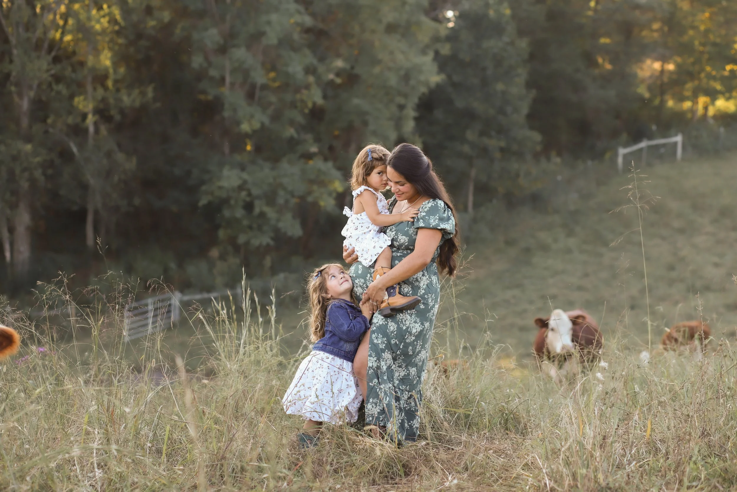 A woman with two young girls in a grassy field with cows and trees in the background during sunset.
