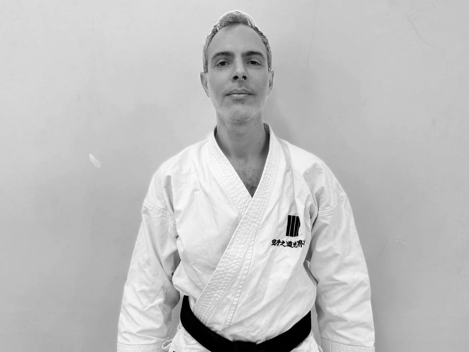 A man in a martial arts gi with a black belt standing against a plain wall.