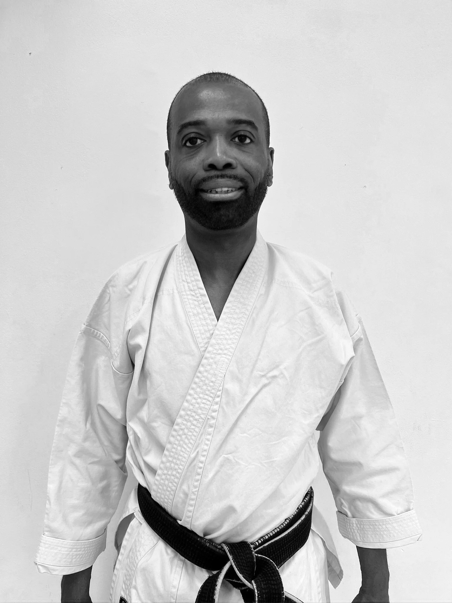A man in a karate gi with a black belt standing in front of a plain wall.