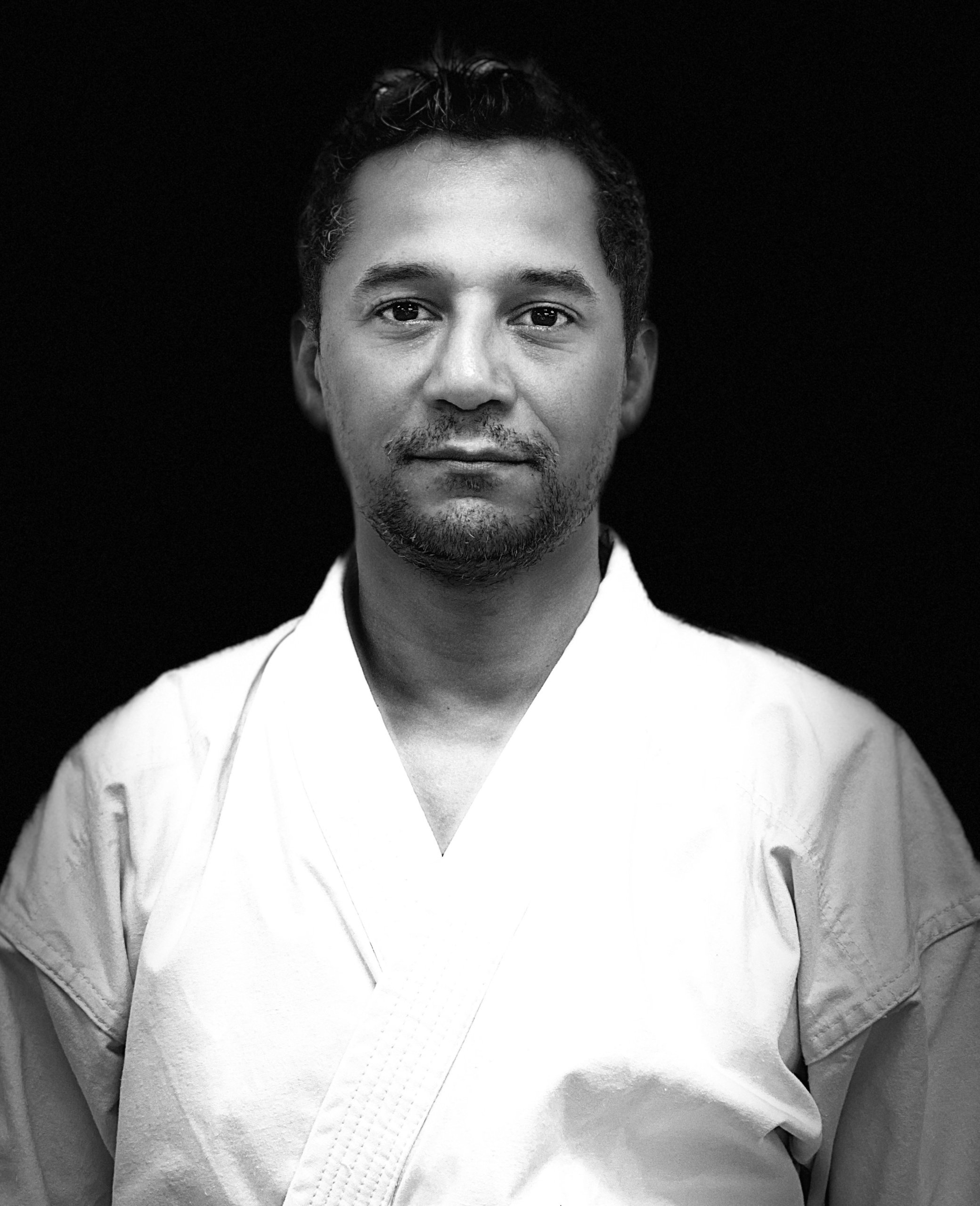 A black and white portrait of a man in a martial arts uniform, with a neutral expression, against a dark background.