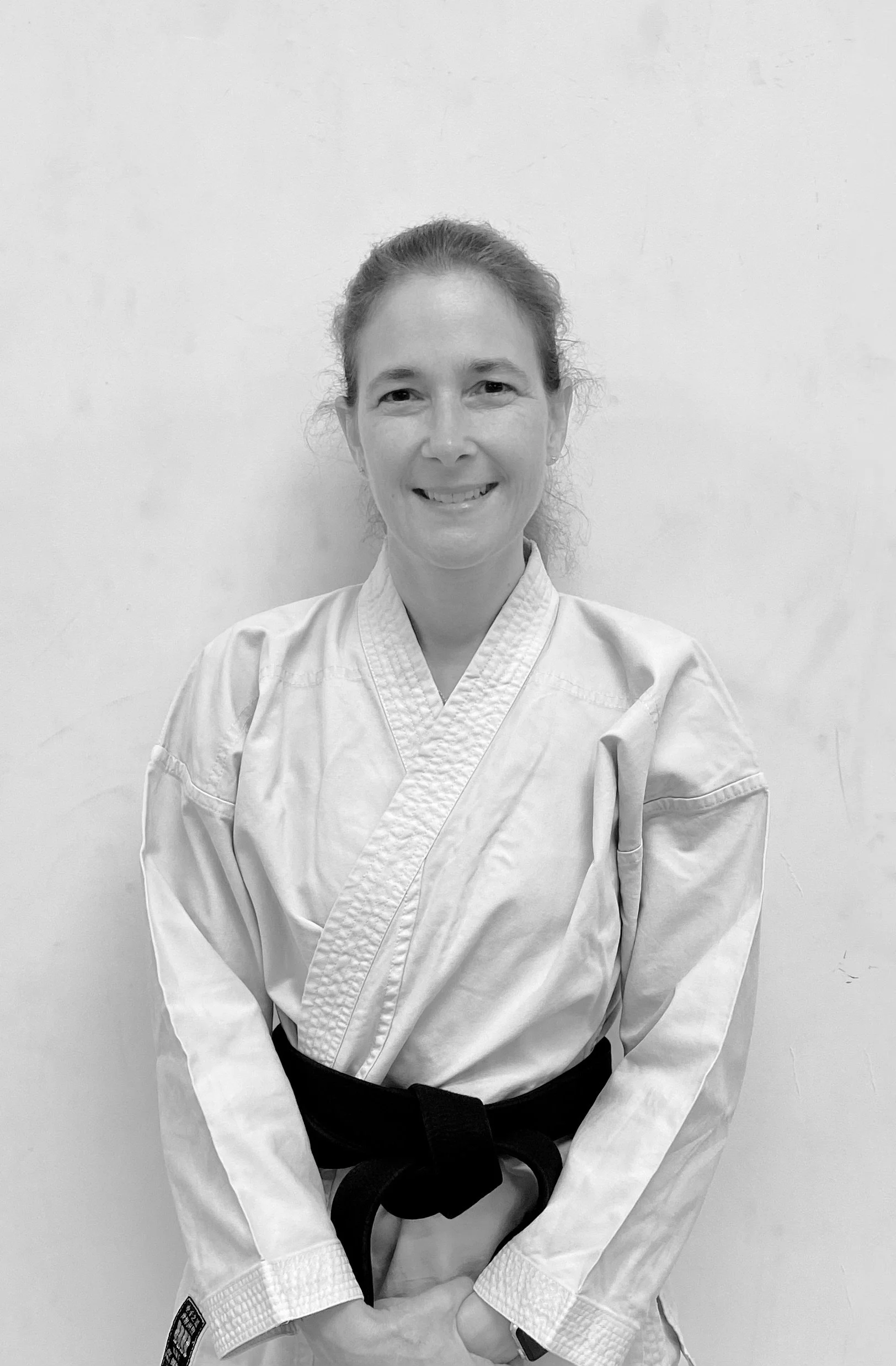 A woman wearing a traditional martial arts uniform with a black belt, smiling at the camera, standing against a plain wall.