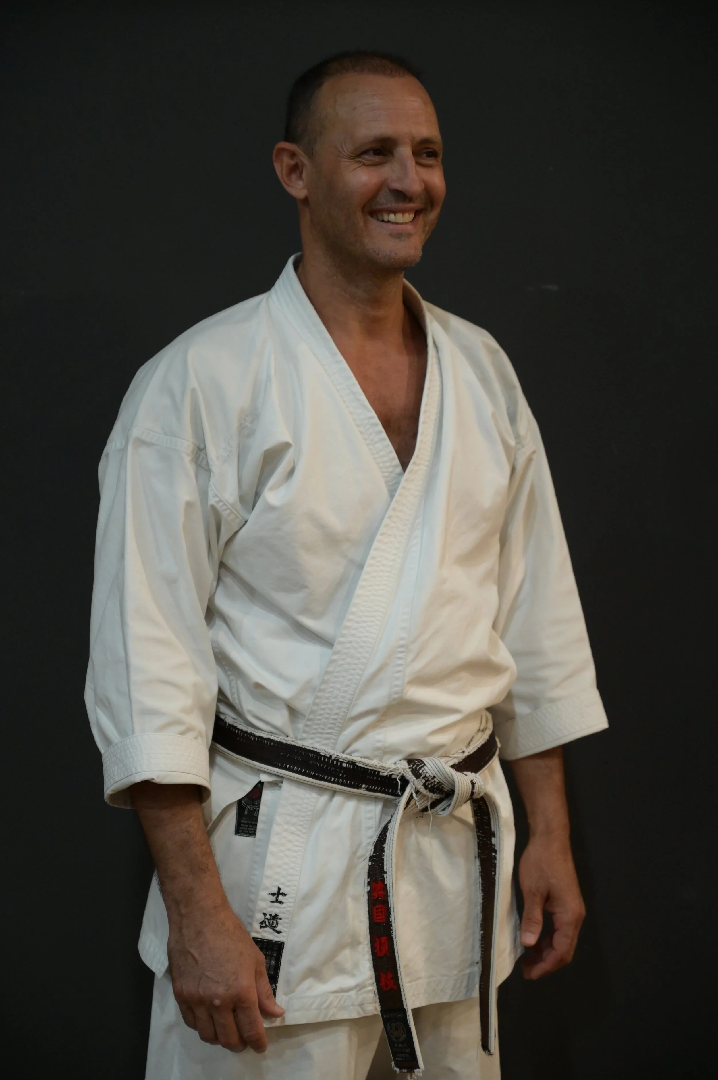 A man in a white karate gi with a black belt, smiling and standing against a dark background.